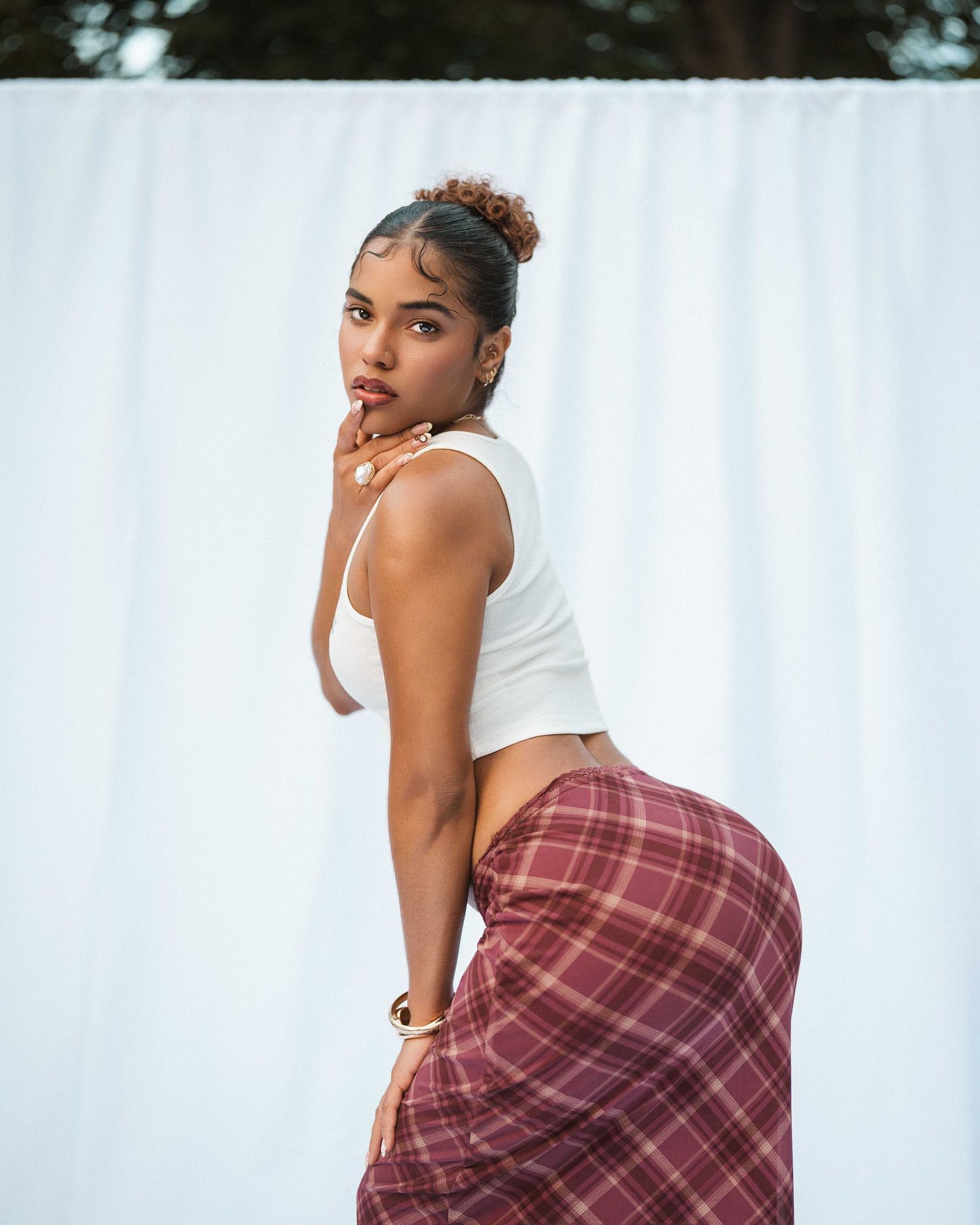 Woman in white crop top and plaid skirt poses outdoors against white fabric backdrop.