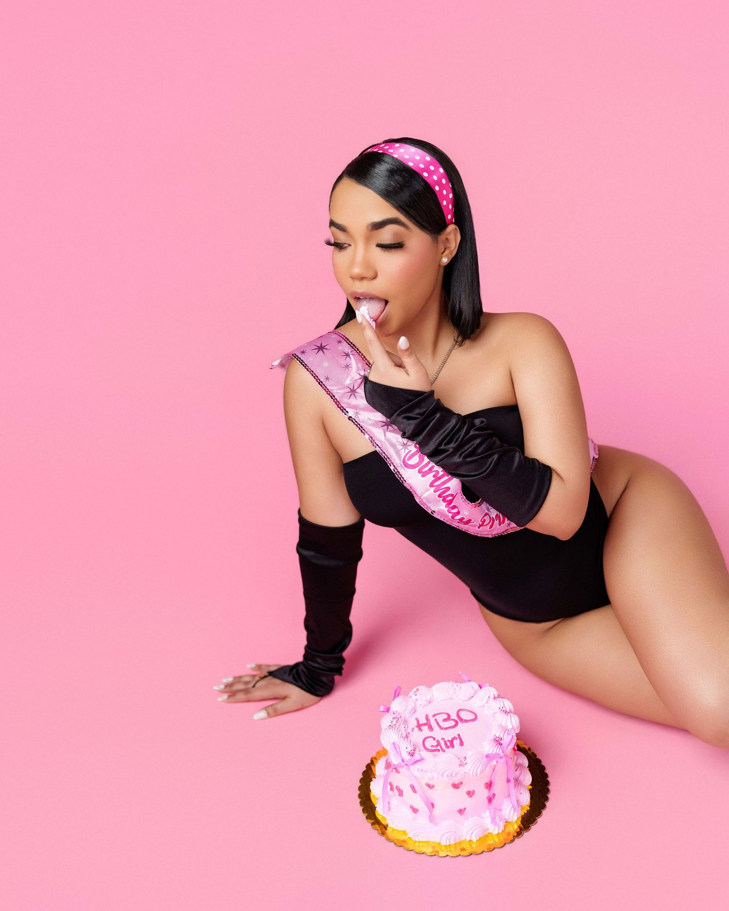 Woman in black bodysuit with birthday sash poses with pink cake on pink background.