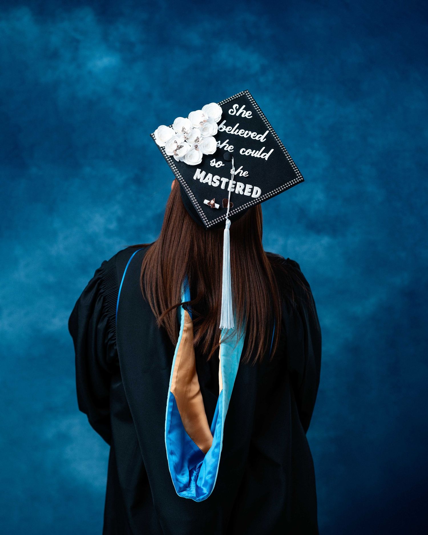 Graduate in black gown and mortarboard cap reading She Believed She Could So She Mastered, decorated with white flowers.