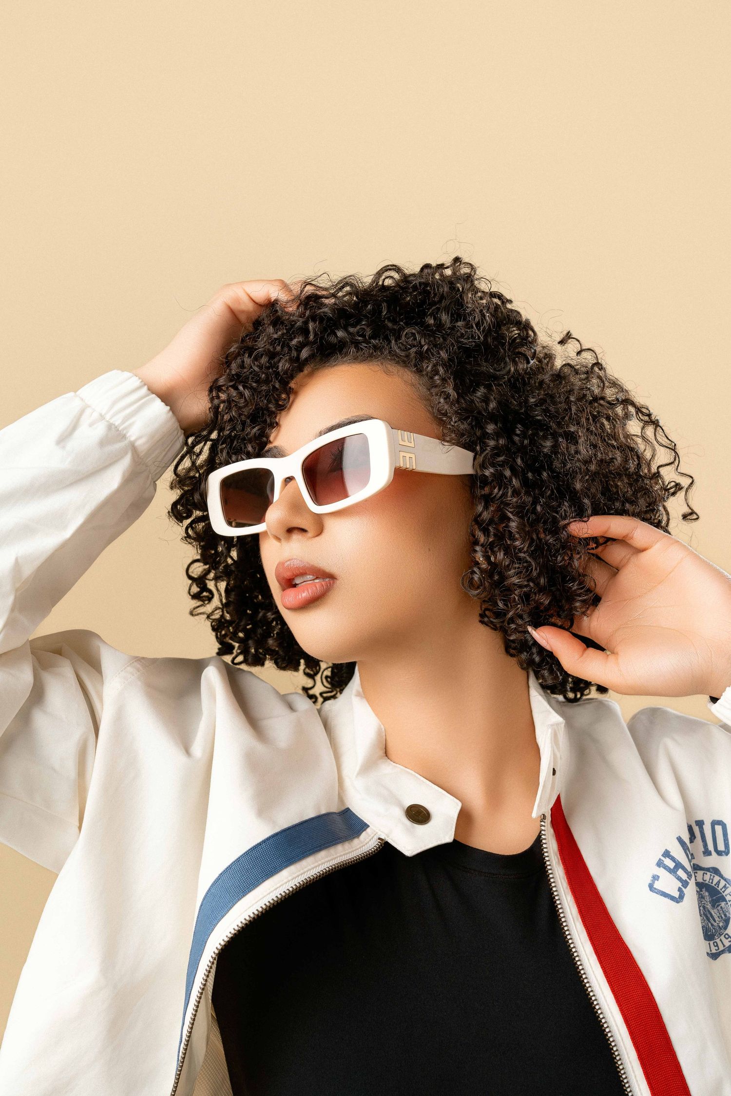 Woman with curly hair wearing white sunglasses and a varsity jacket on a beige background.