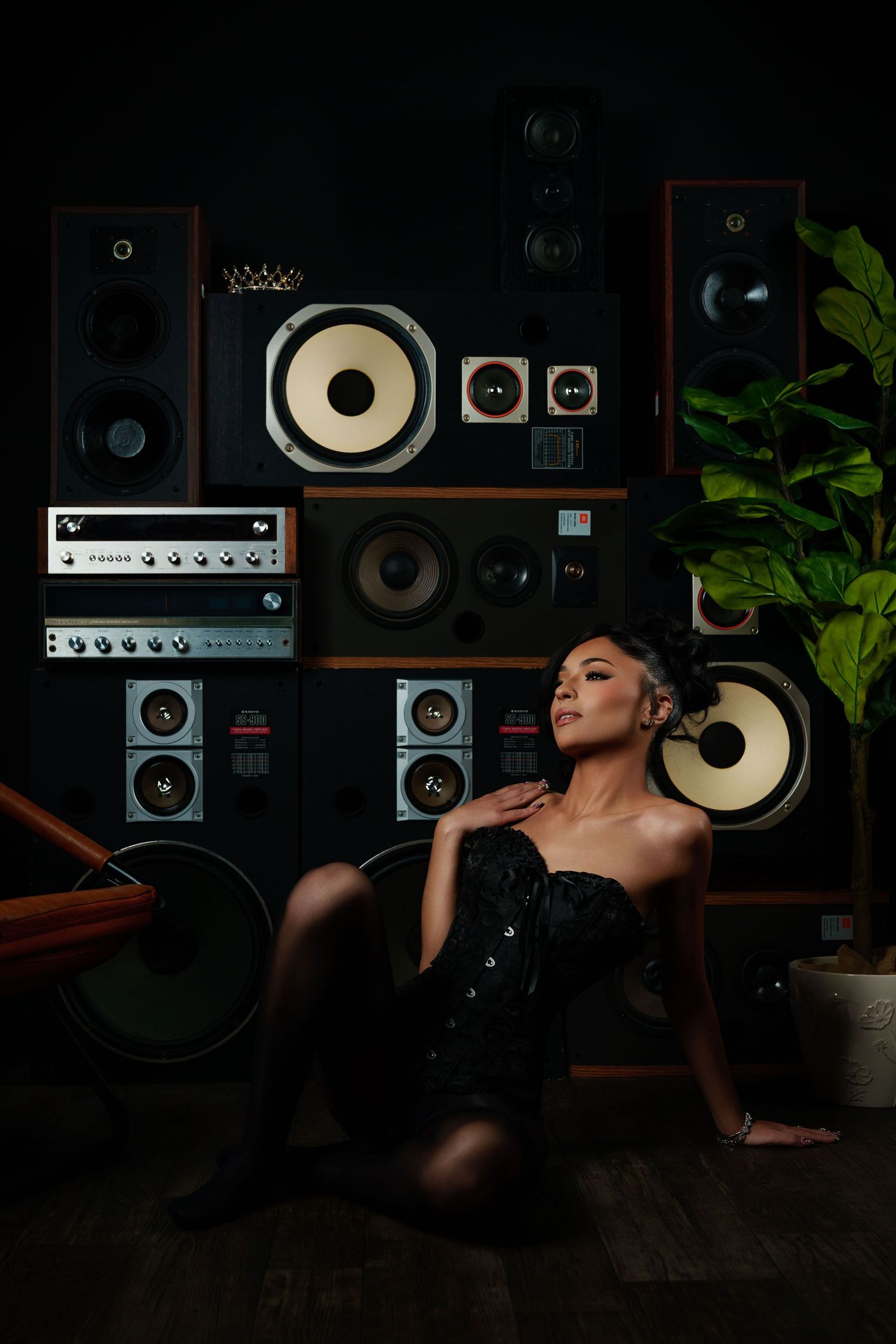 Woman in black corset sits on floor before wall of vintage audio speakers and plant.