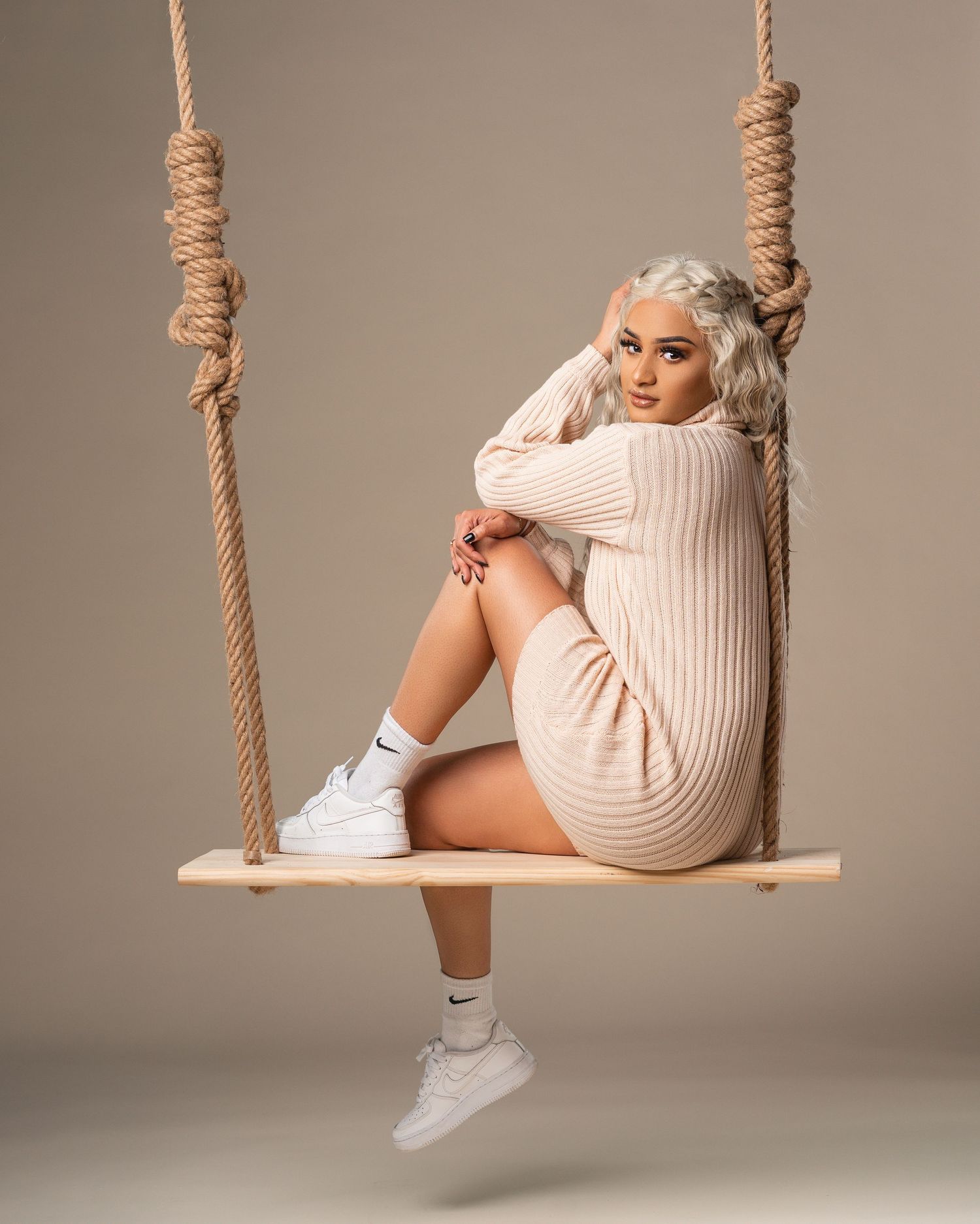 Woman in beige knit dress and white Nike sneakers sitting on a wooden rope swing in a studio.