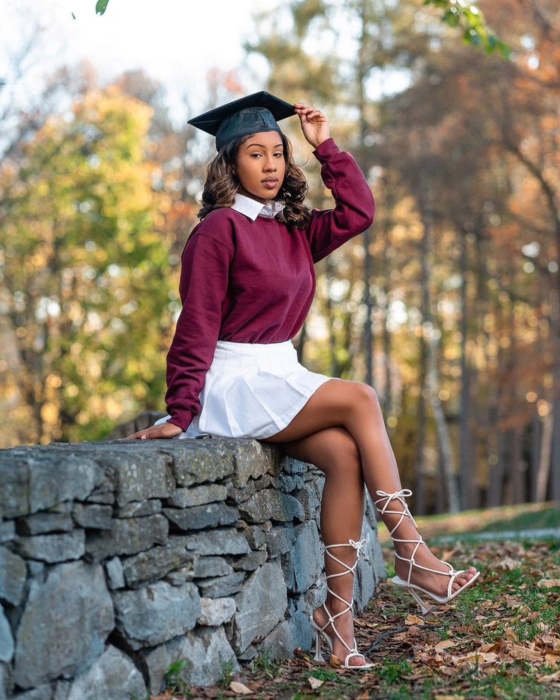 A young woman in a maroon sweater, white skirt, and graduation cap poses on a stone wall outdoors in autumn.