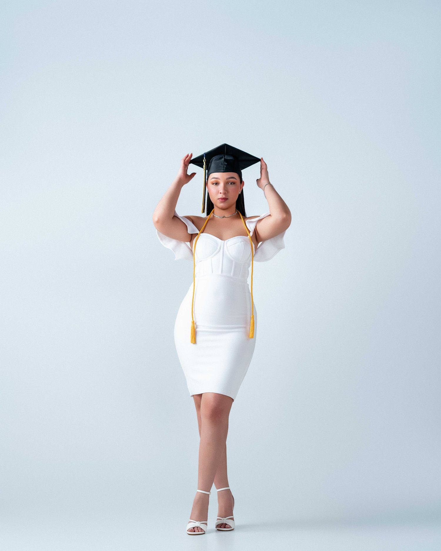 Woman in white dress and graduation cap posing for a professional graduation photo on a light background.