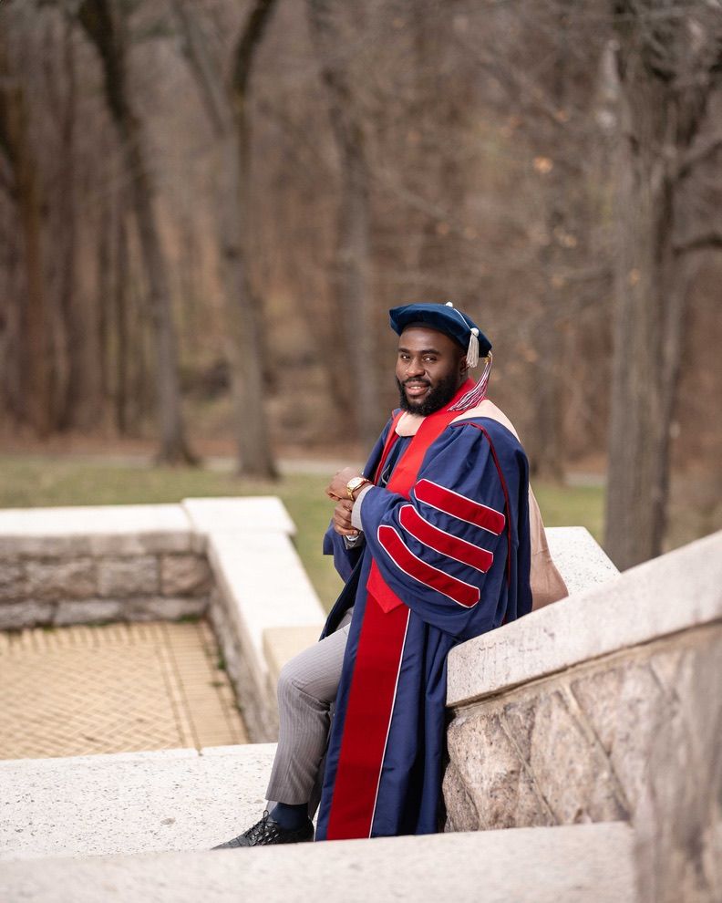 Man in doctoral graduation regalia smiles while seated on stone steps outdoors among bare trees.