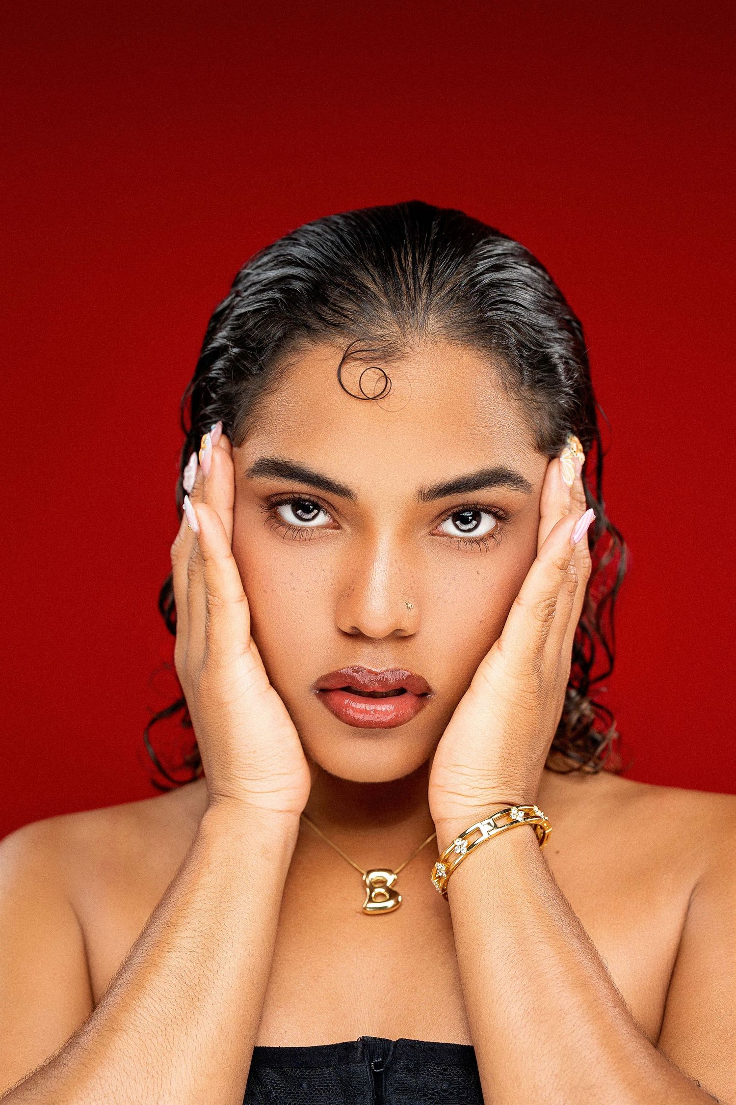 Close-up portrait of a woman with gold jewelry holding her face against a red background.