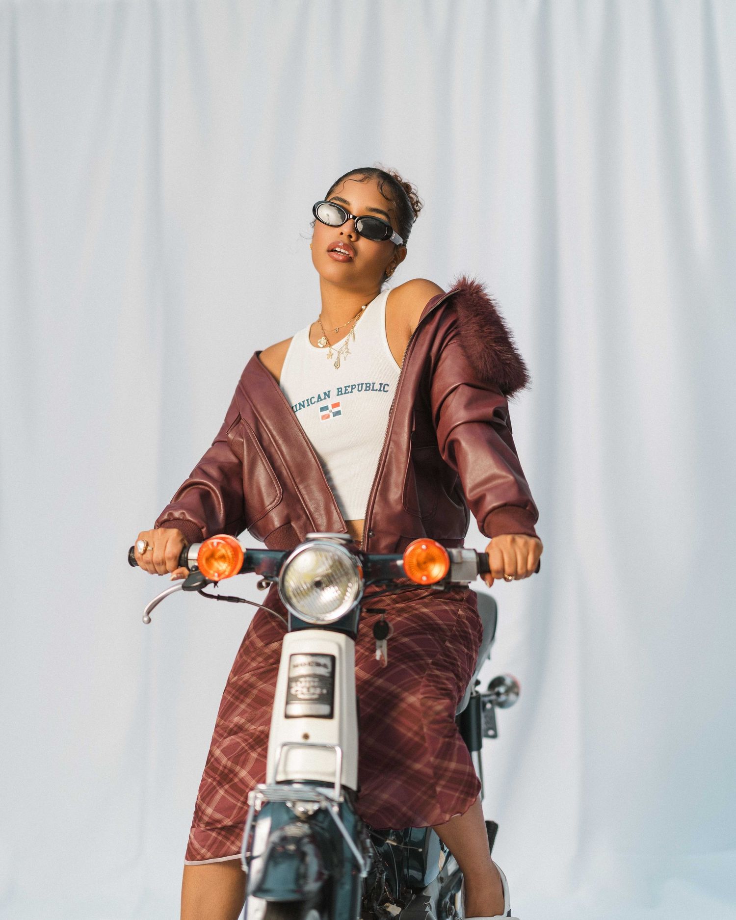 Woman in burgundy fur jacket and plaid skirt poses on vintage motorcycle against white backdrop.