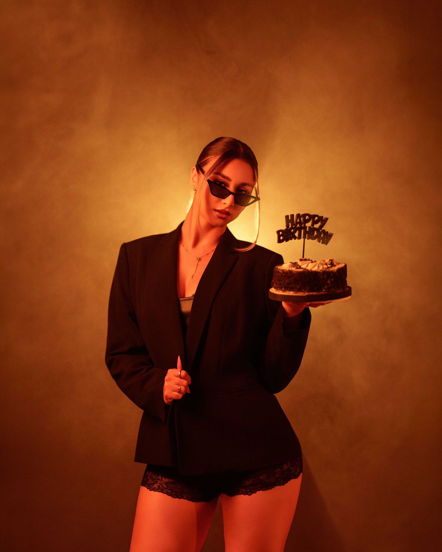 Woman in black blazer and sunglasses holding a Happy Birthday cake against a warm golden-orange background.