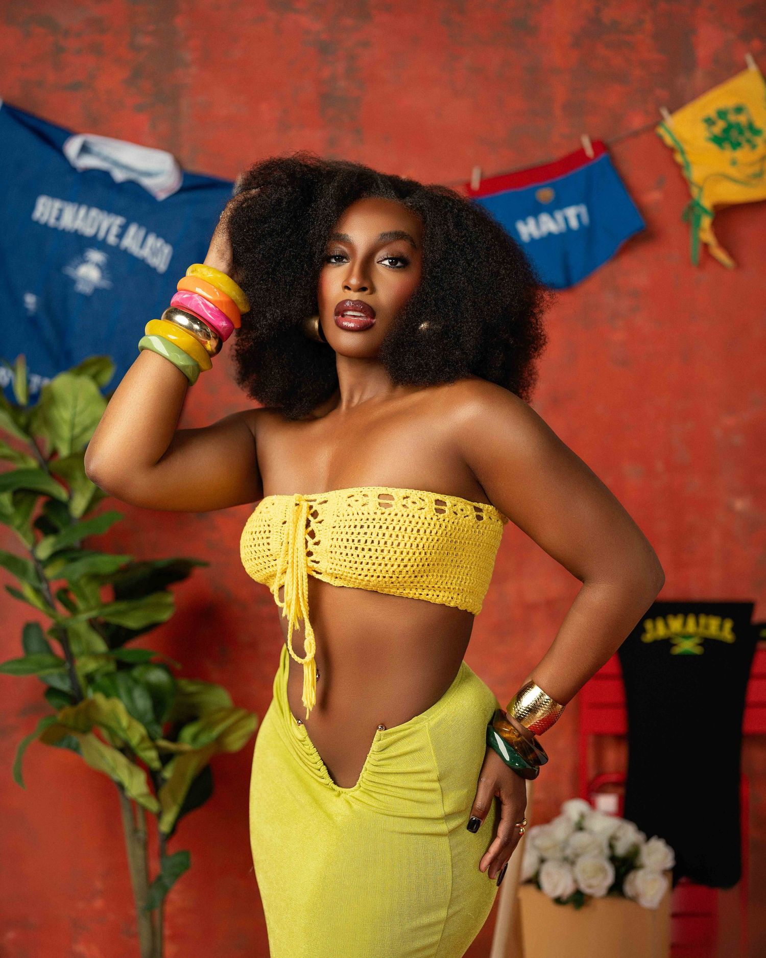 Woman in yellow crochet crop top and skirt poses with colorful bangles against red wall.
