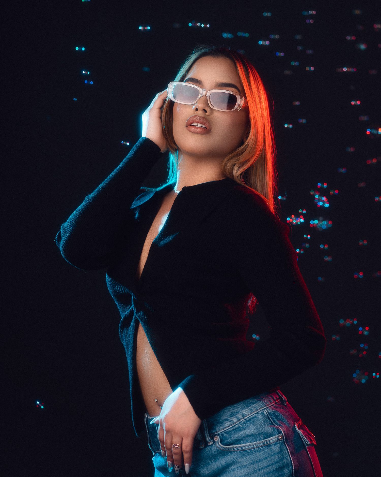 Woman in black top and jeans wearing white sunglasses under colorful neon studio lighting.