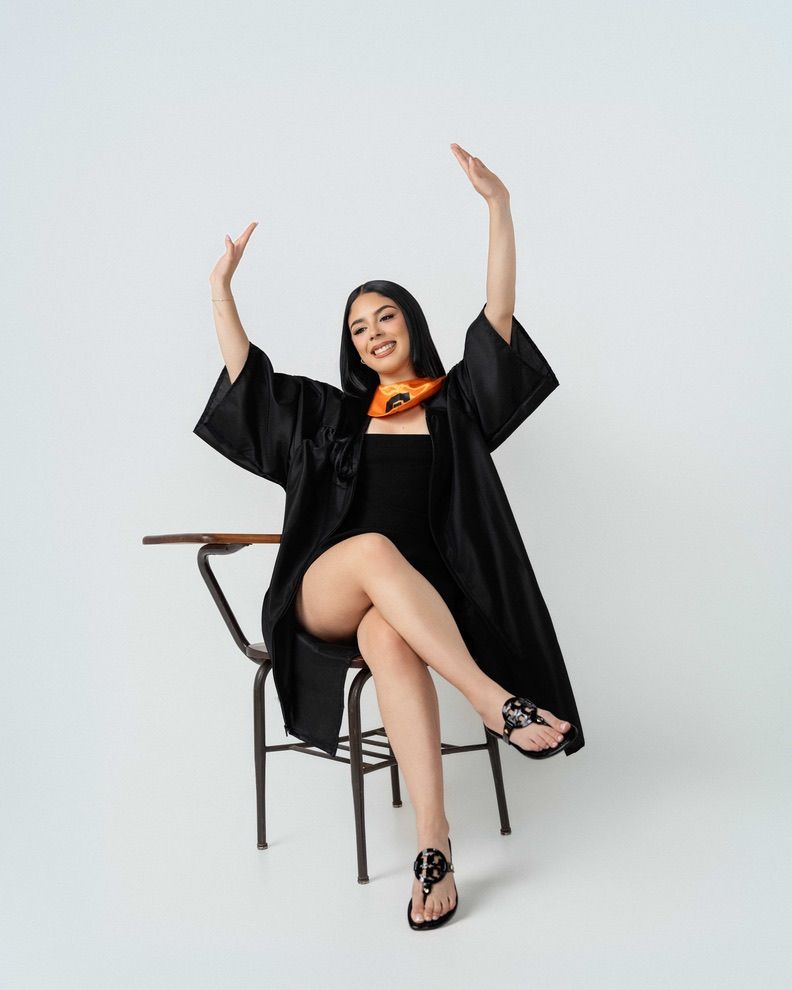 Woman in black graduation robe sits on chair with arms raised, wearing sandals and colorful scarf, white background.