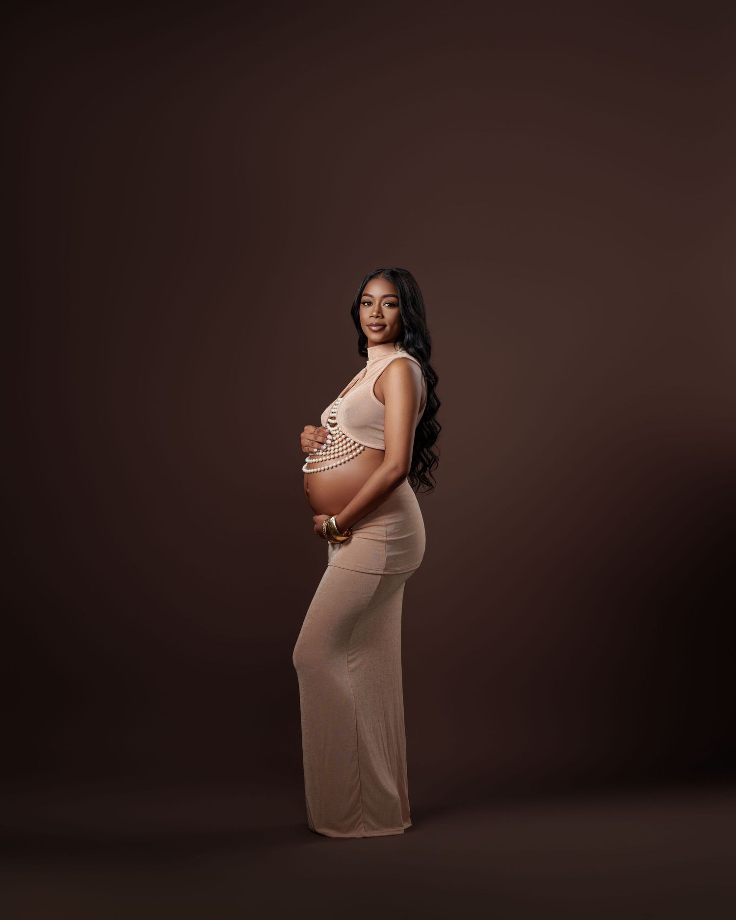 Pregnant woman in beige form-fitting dress cradling her bump against a dark brown background.