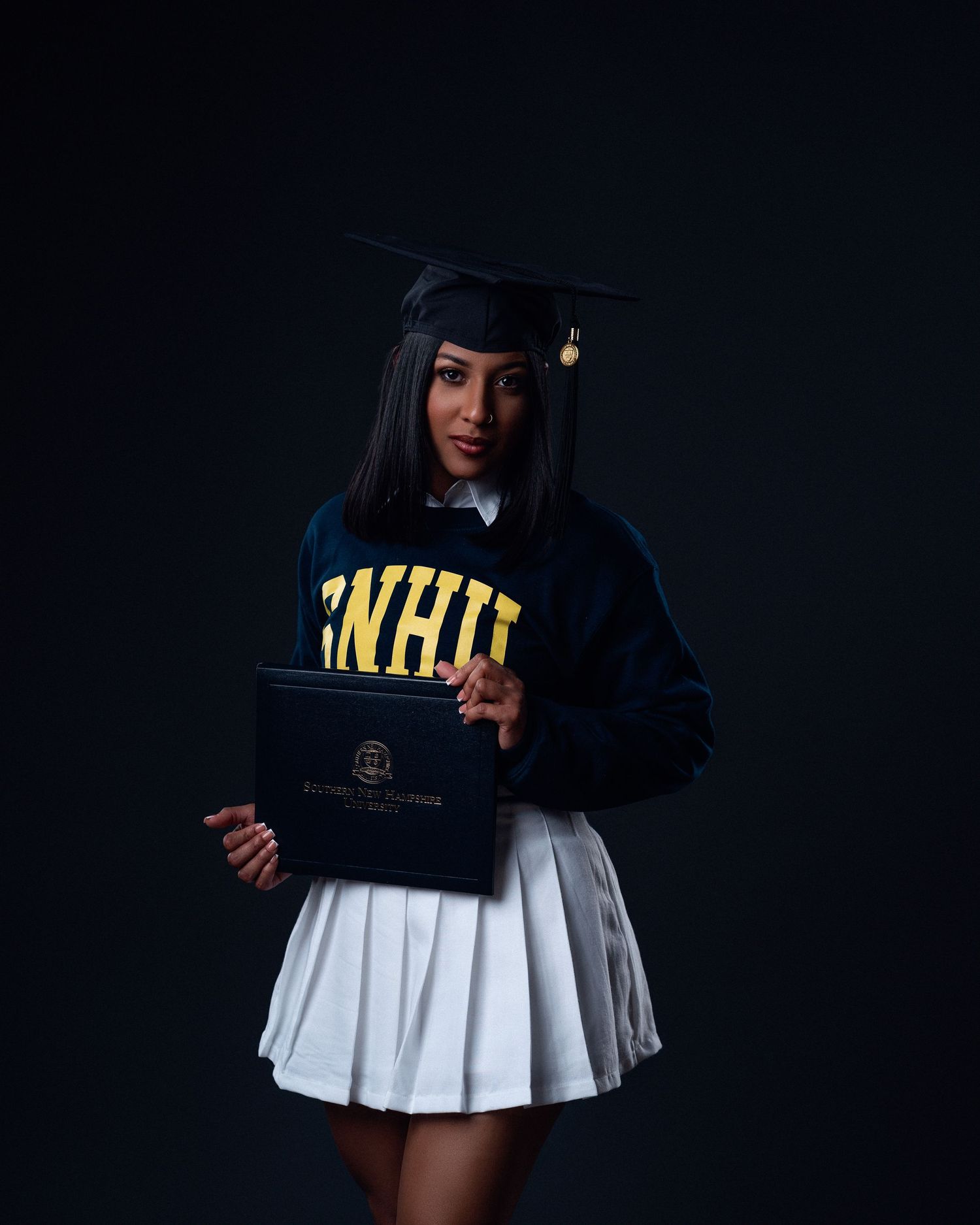 SNHU graduate in navy college sweater and white pleated skirt holds diploma wearing black graduation cap.