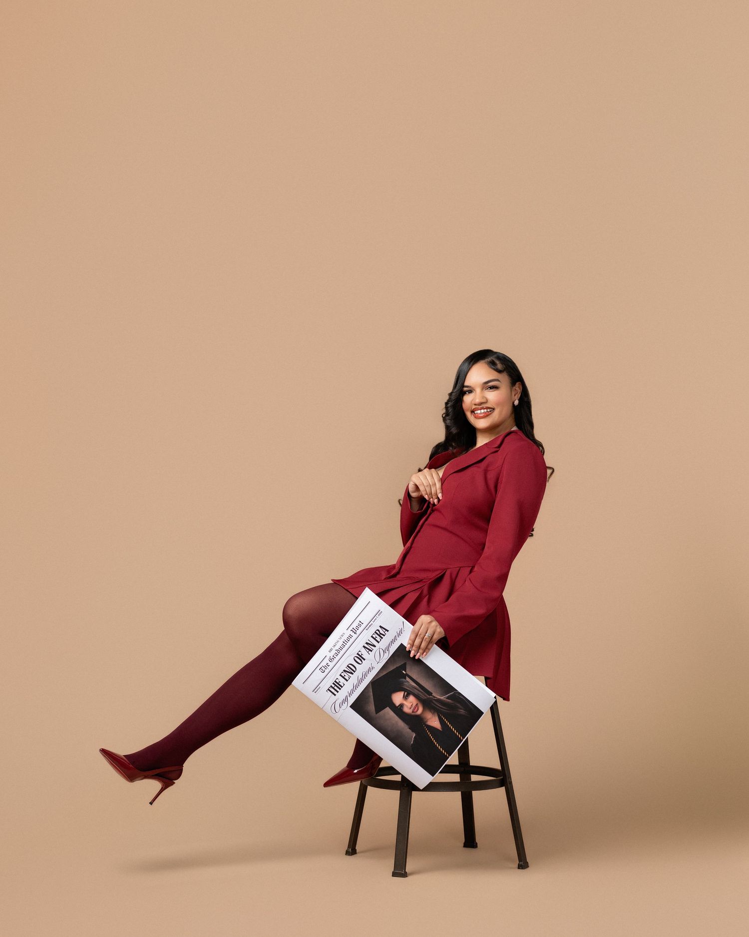 Woman in red dress and boots sits on stool holding a magazine, posing confidently against a beige background.