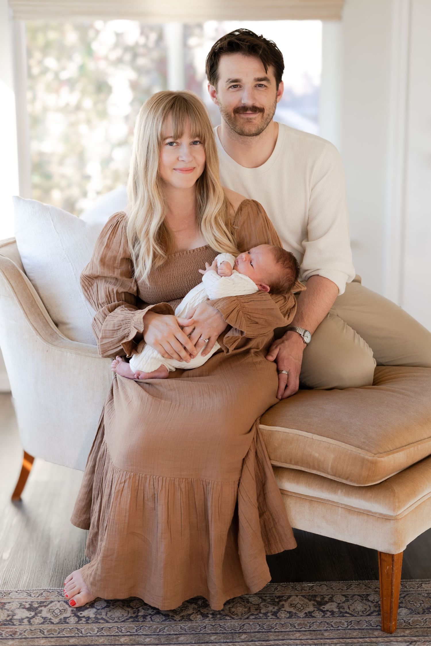 Indoor family portraits on a beige couch showing parents with their newborn baby.
