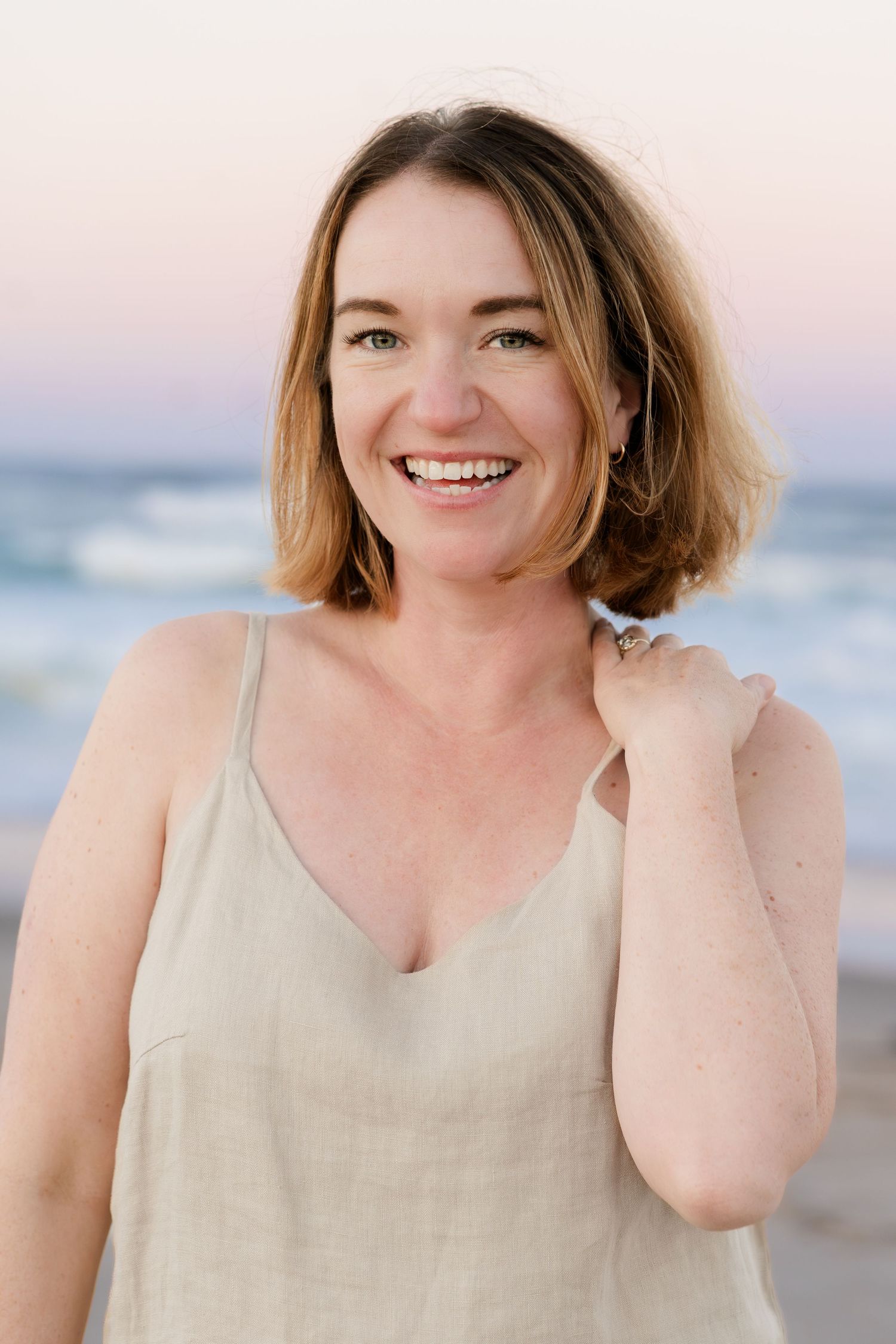 Person in a beige sleeveless top smiling joyfully at the beach during golden hour.