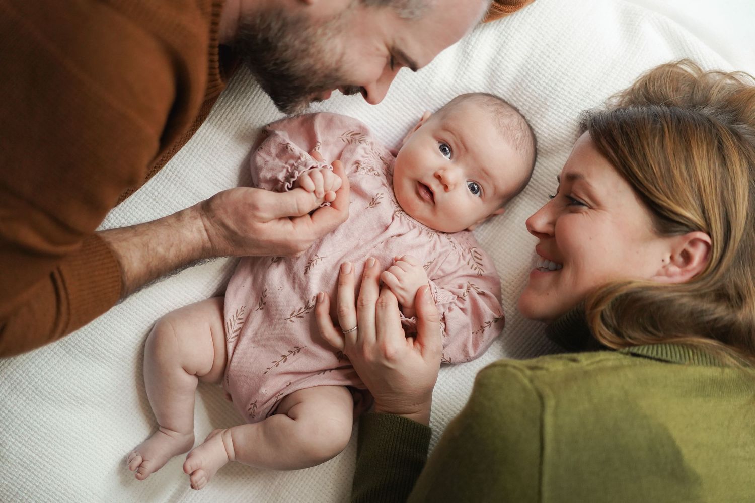 Family sharing intimate moment with baby wearing pink romper on white bedding.