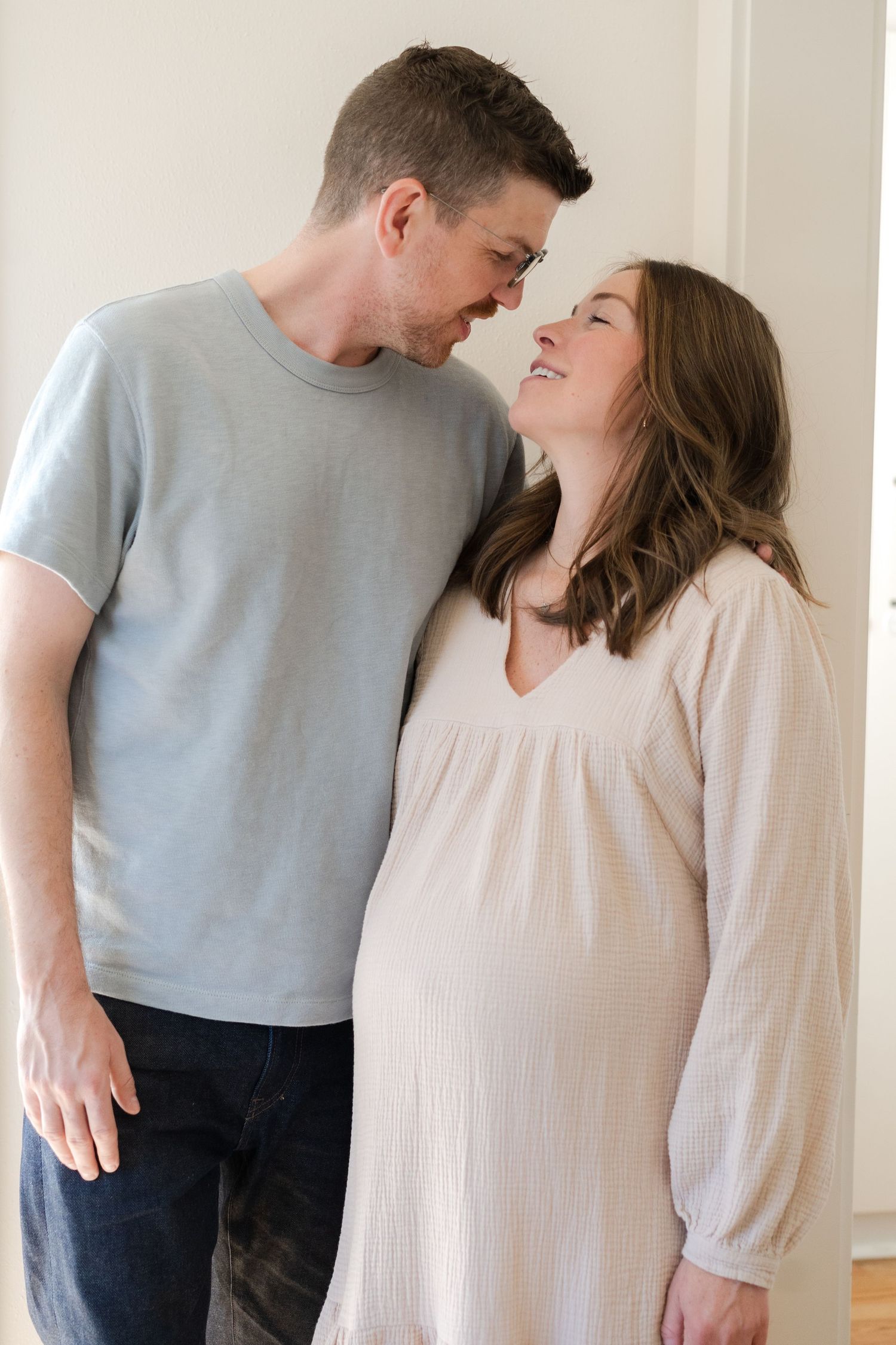 A couple shares an intimate moment in casual clothing against a white wall.