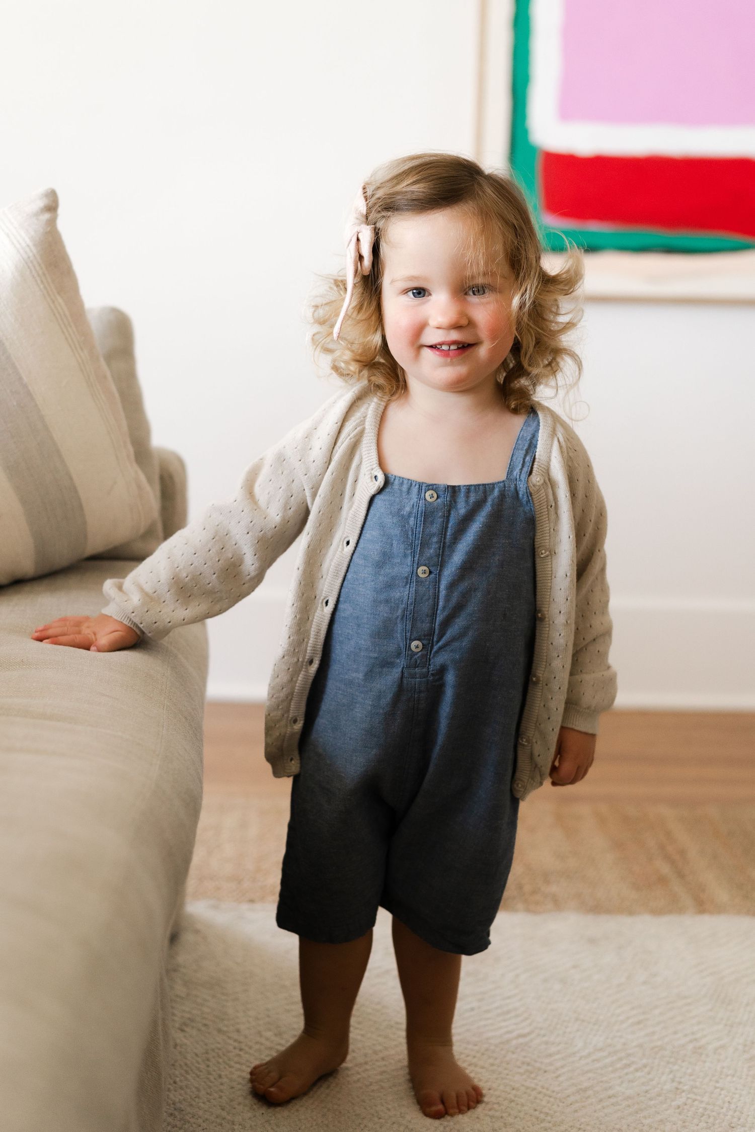 Toddler standing barefoot in denim overalls against a light colored wall.