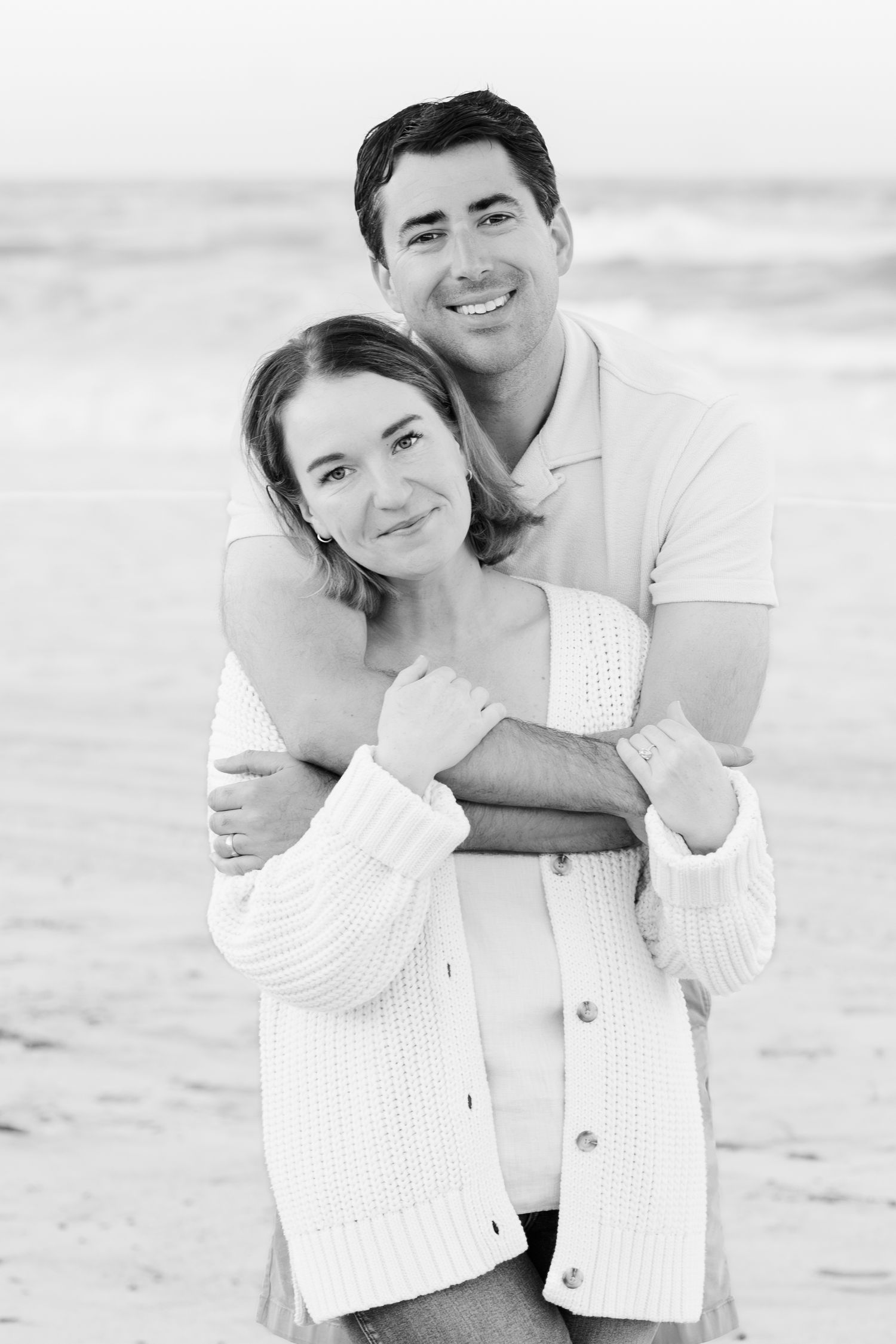 A couple embracing on the beach in black and white, wearing casual attire.