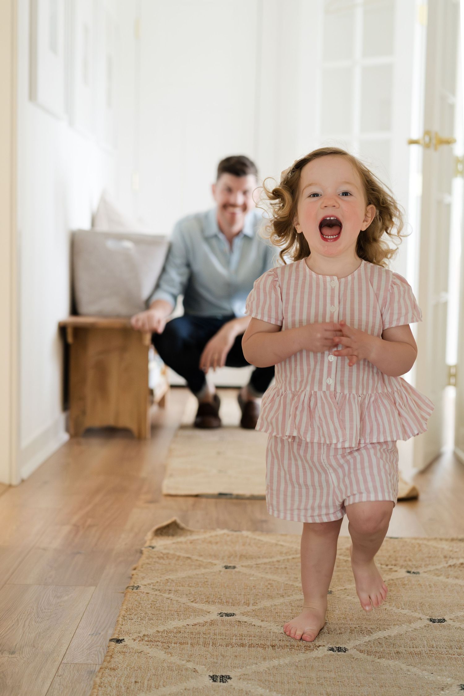 A toddler in pink stripes runs excitedly down a sunlit hallway.
