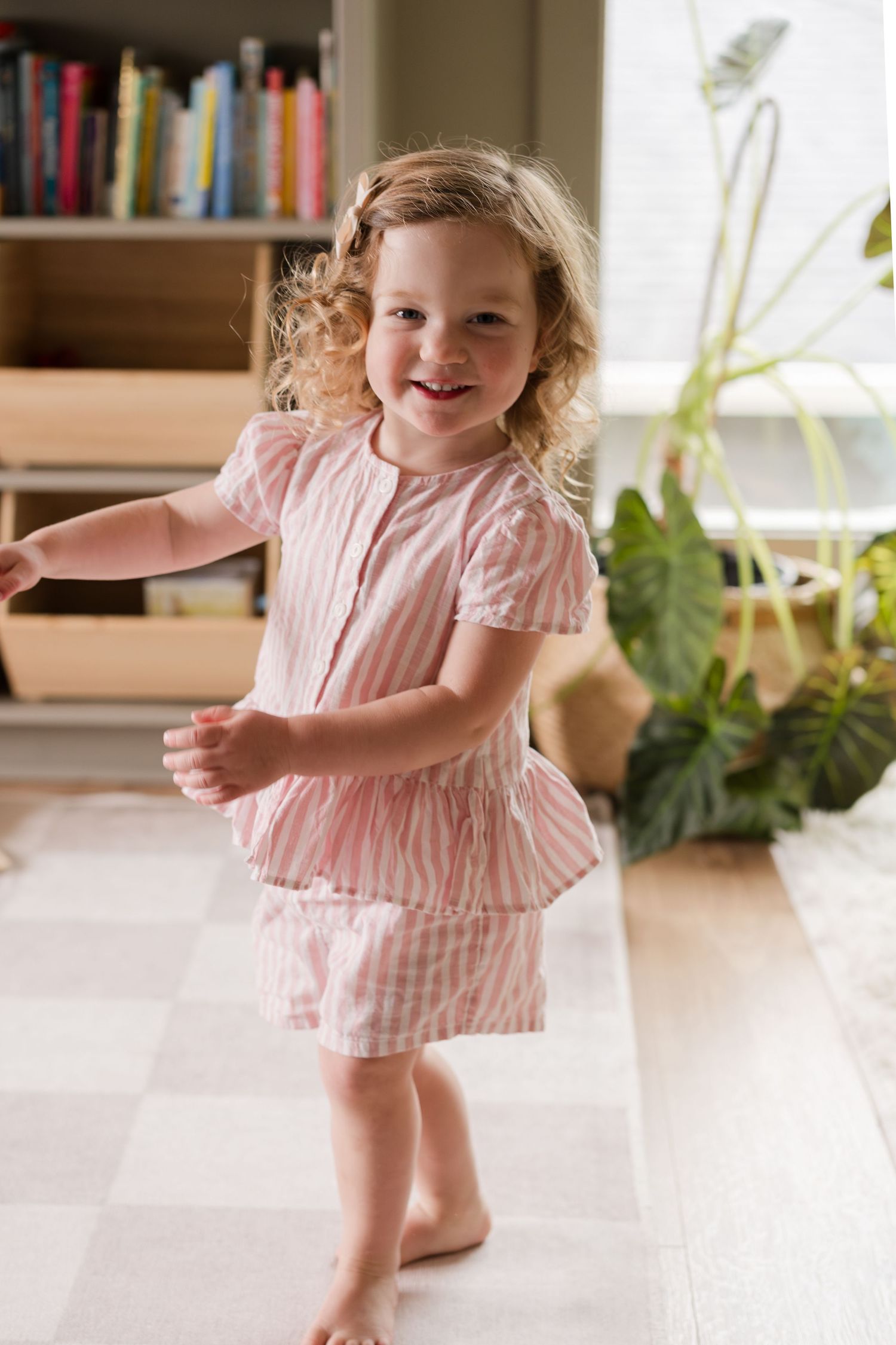 A toddler in pink twirls and dances in a sunlit room with houseplants.