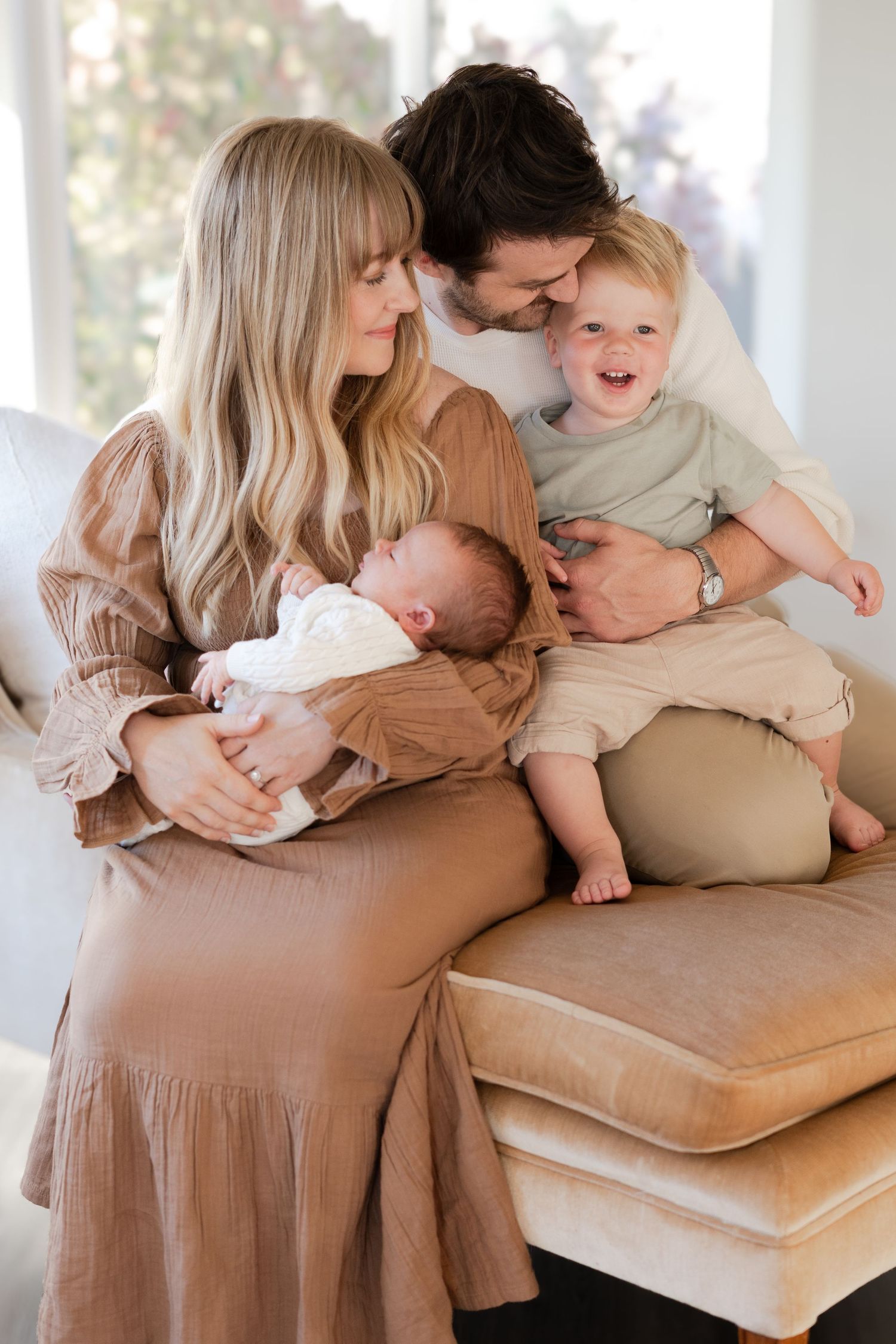 Family members sharing tender cuddles and embraces on a beige couch in a sunlit room.