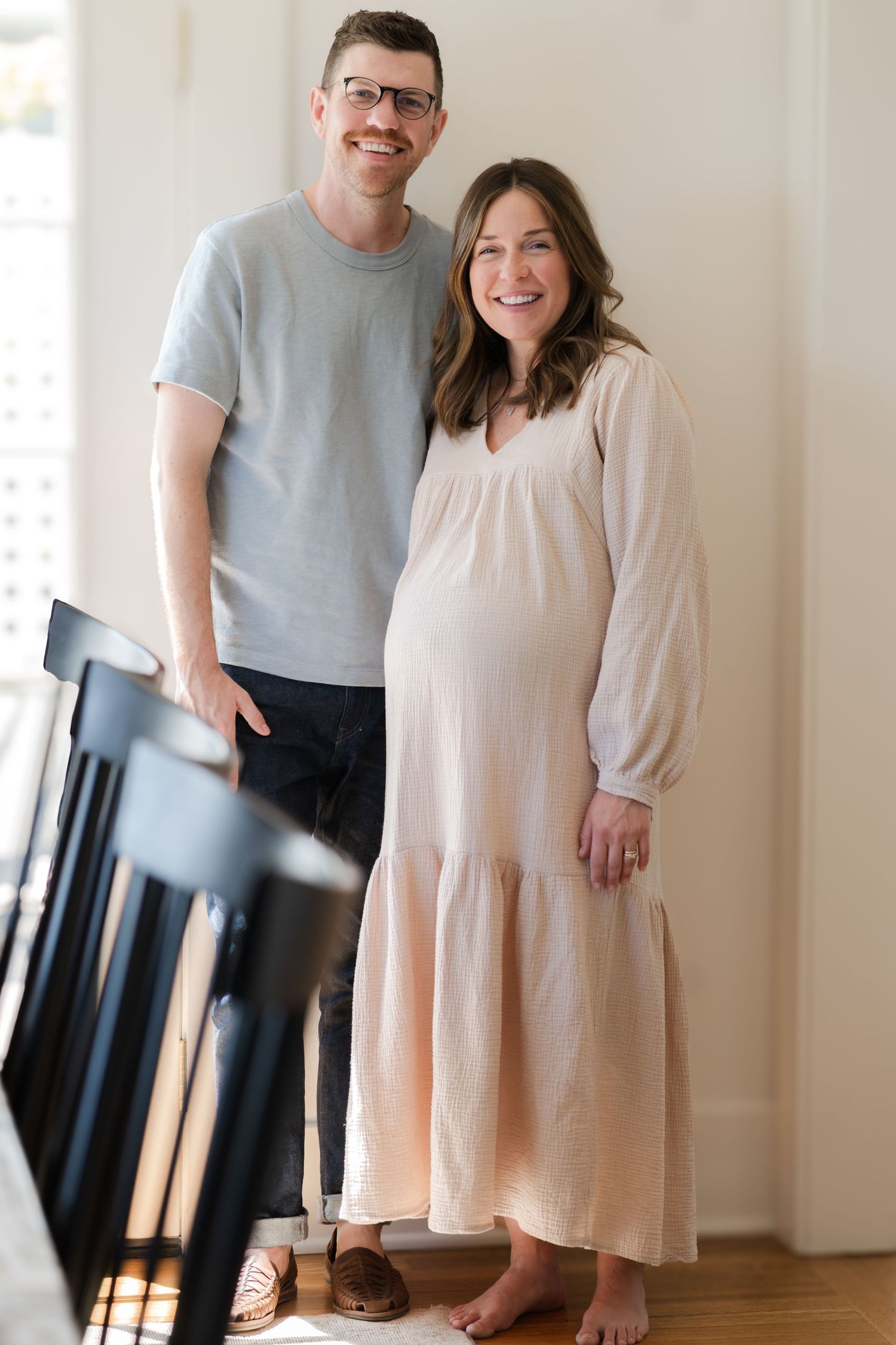 A couple poses together in a sunlit room wearing casual attire.