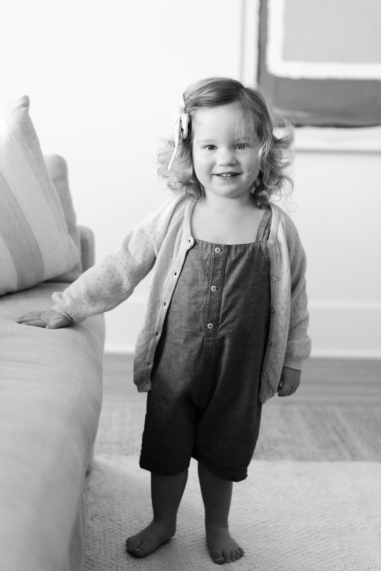 Child in denim overalls stands barefoot on carpet in black and white photo.