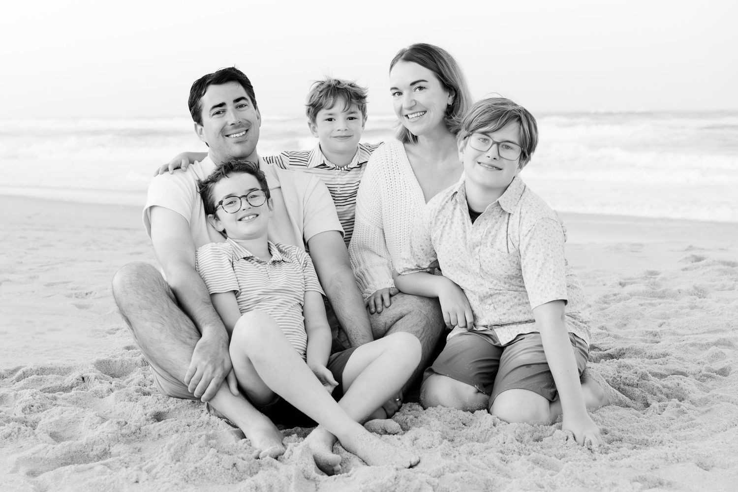Black and white beach portrait of family sitting together in casual poses.