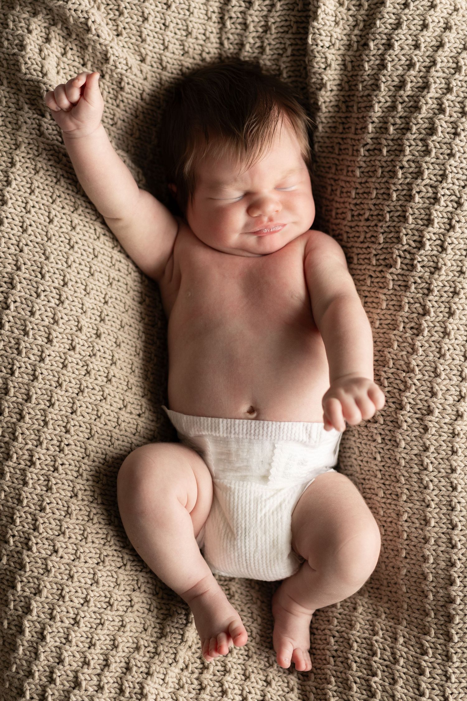 Baby lying on textured beige blanket with arms stretched out.