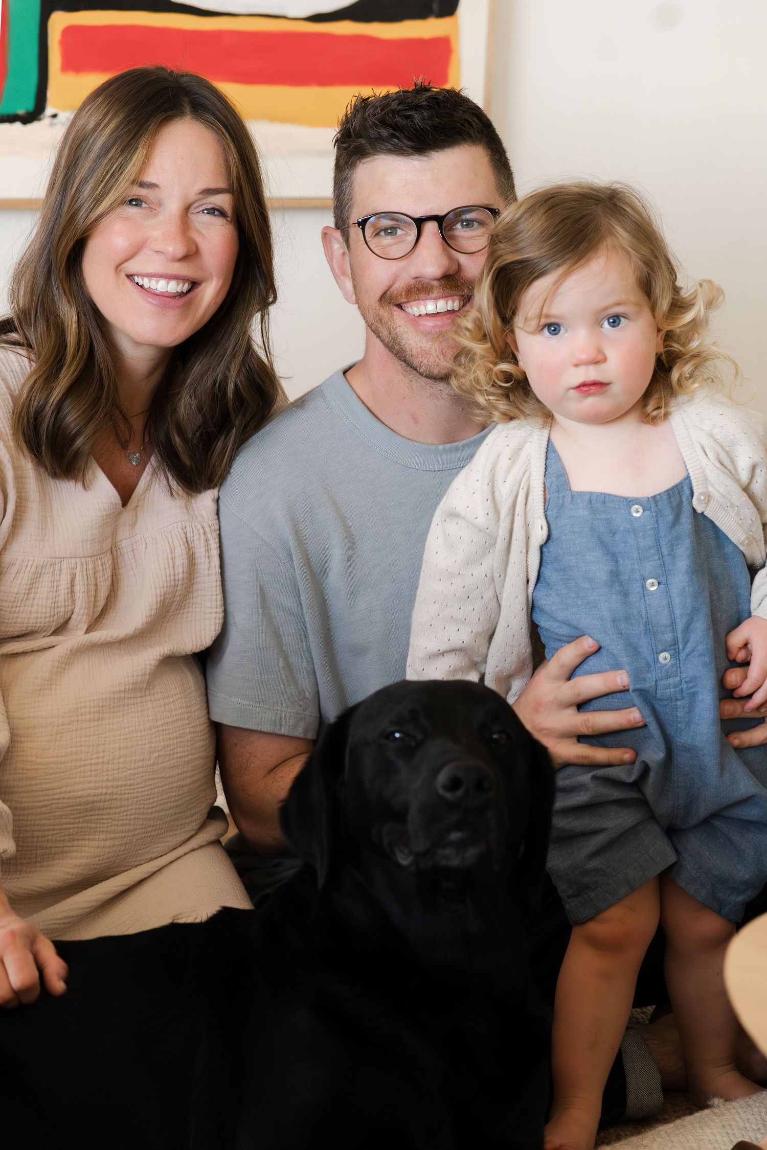 A candid family moment captured on a couch with their pet dog and colorful artwork in background.