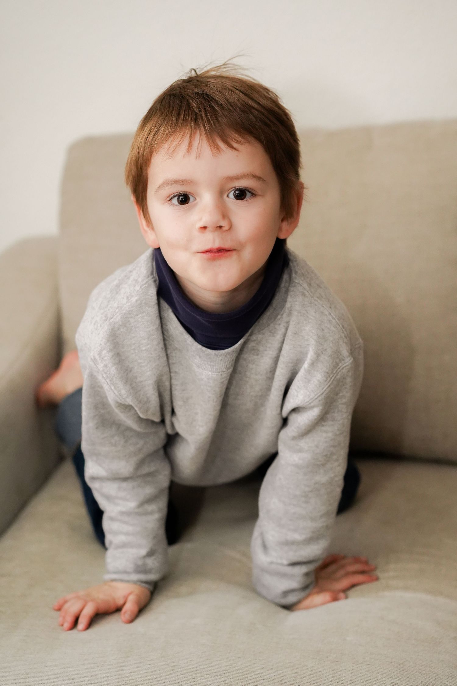 Small child in gray sweatshirt crawling playfully on beige couch.