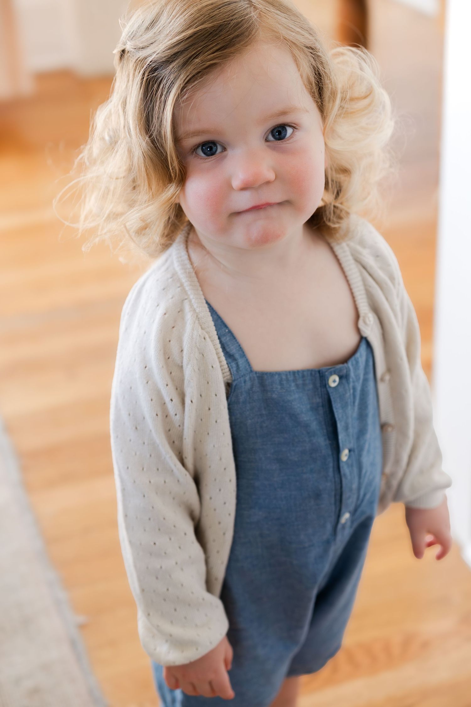 A toddler in denim overalls and cream cardigan stands on hardwood floors.