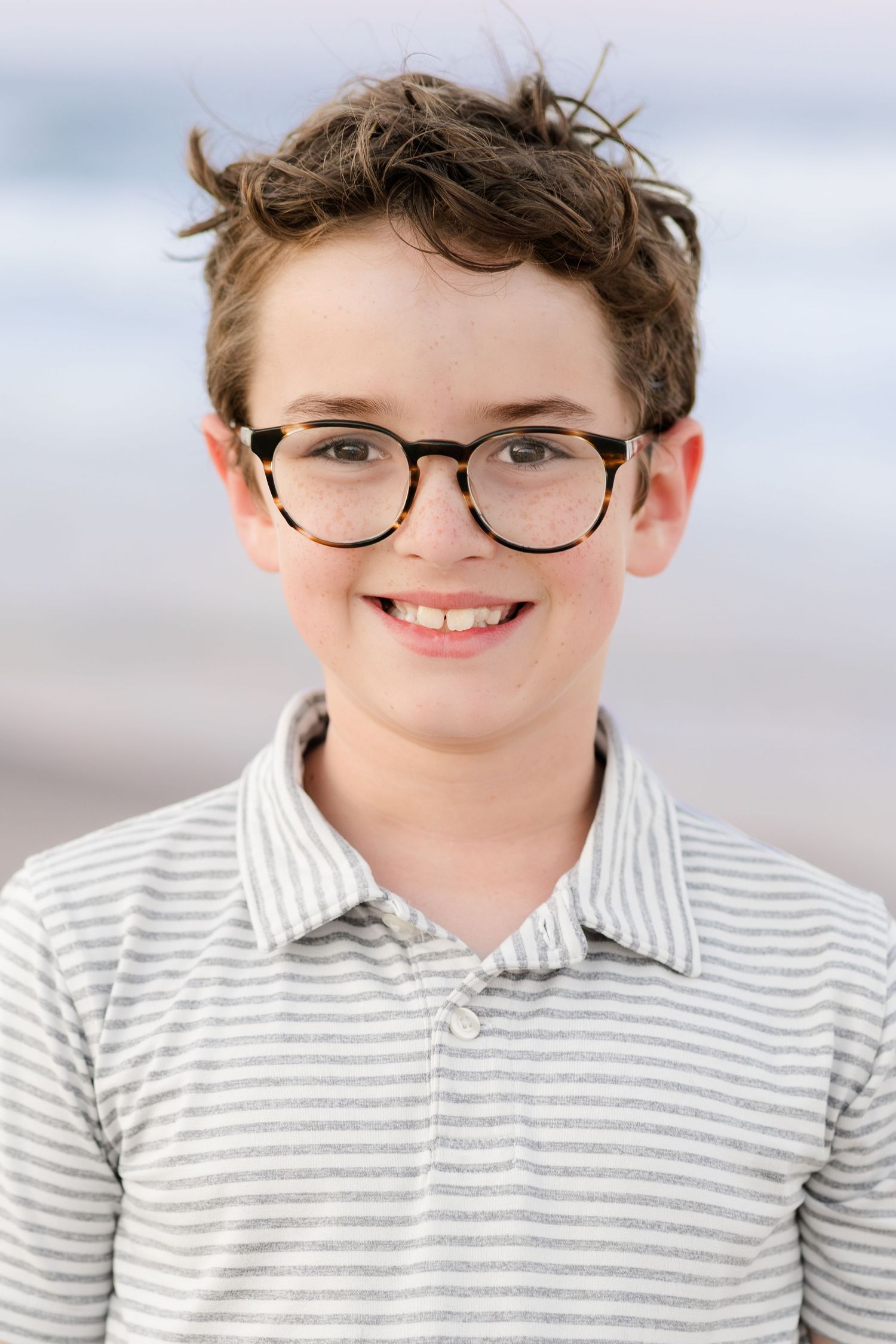 Young person in striped polo shirt and glasses smiles brightly against light background.