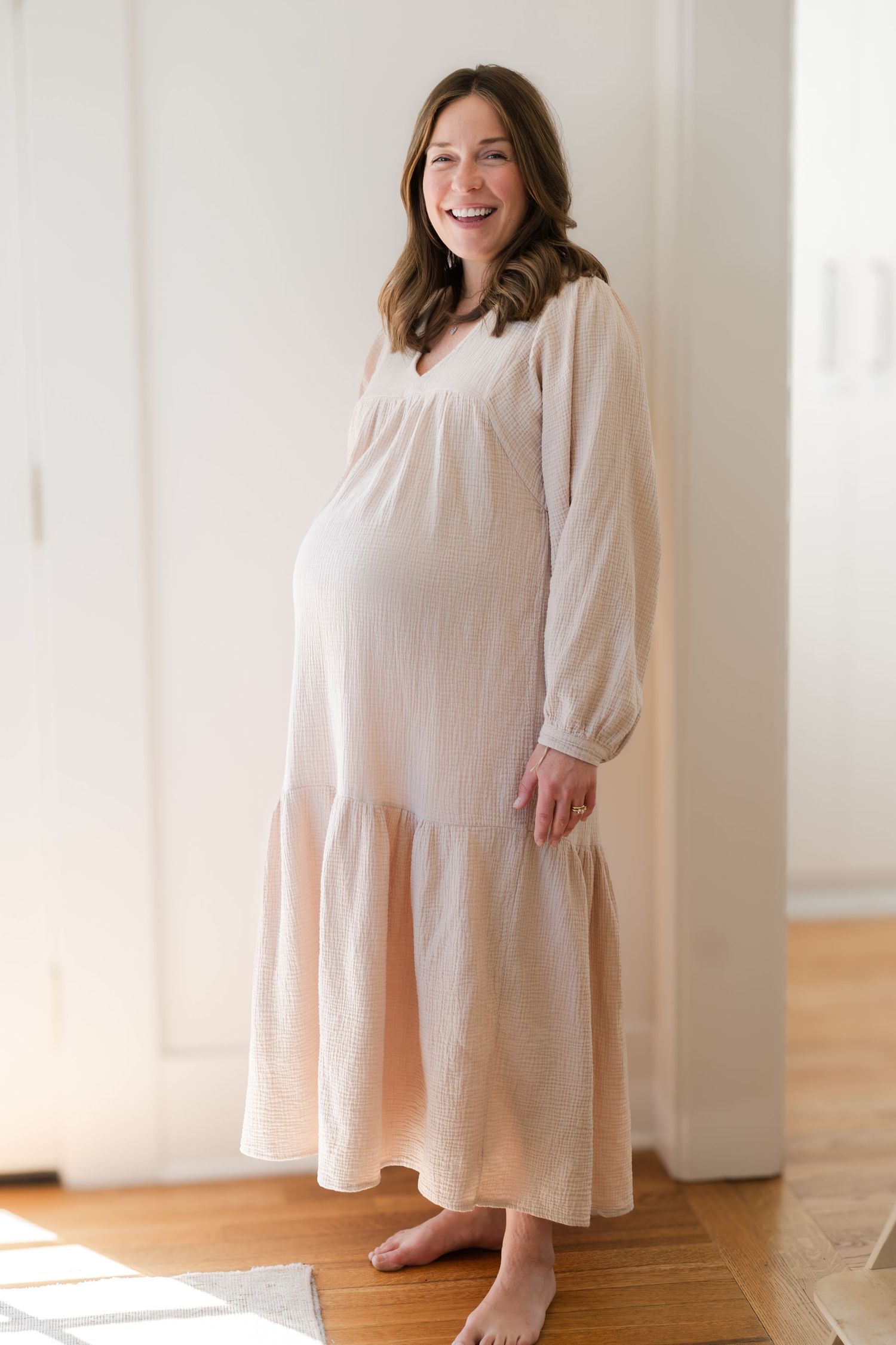 A pregnant person wearing a flowing cream-colored maxi dress stands barefoot in a sunlit room.