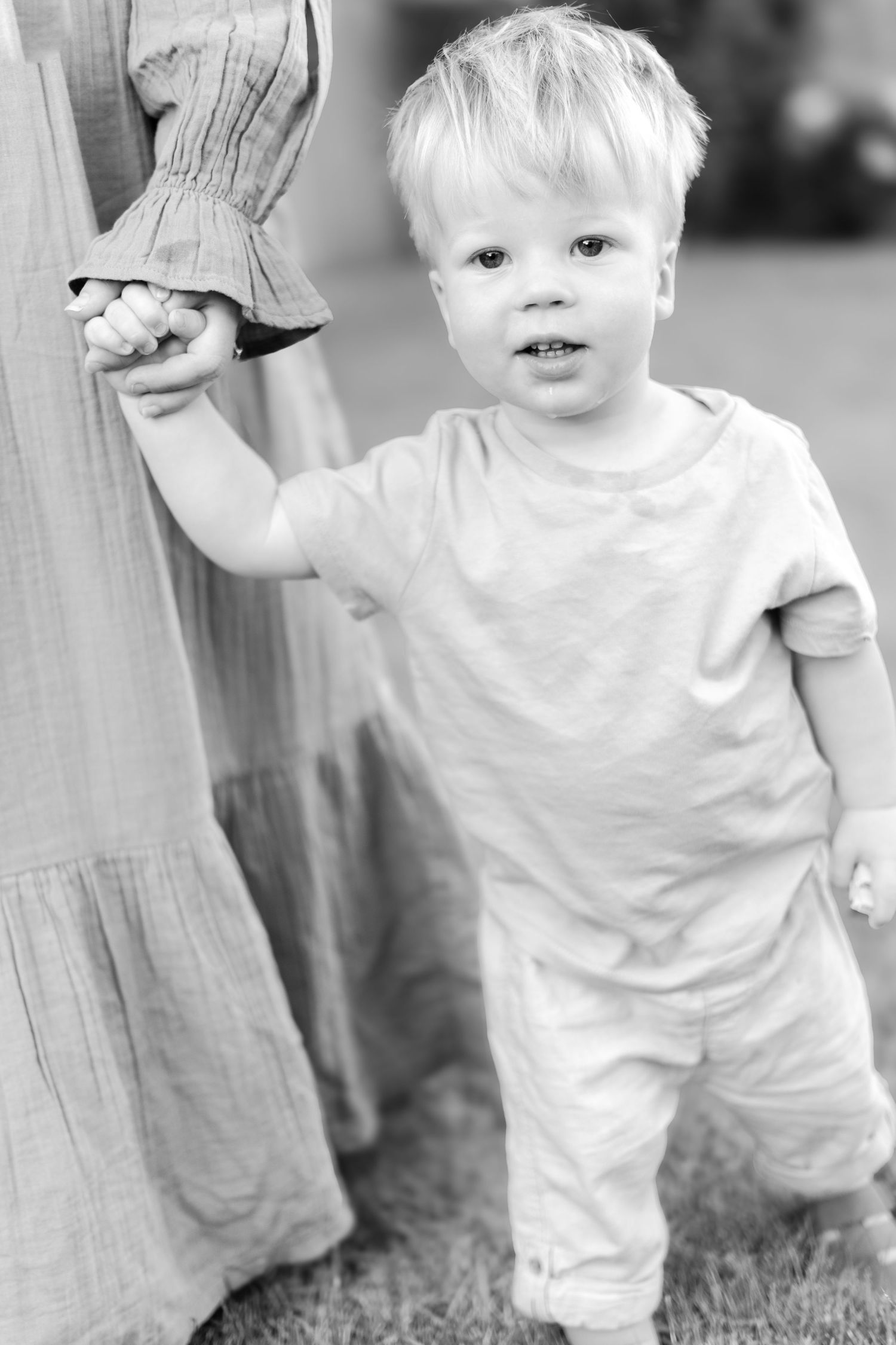 Black and white photo of toddler holding hand of person in flowing dress outdoors.