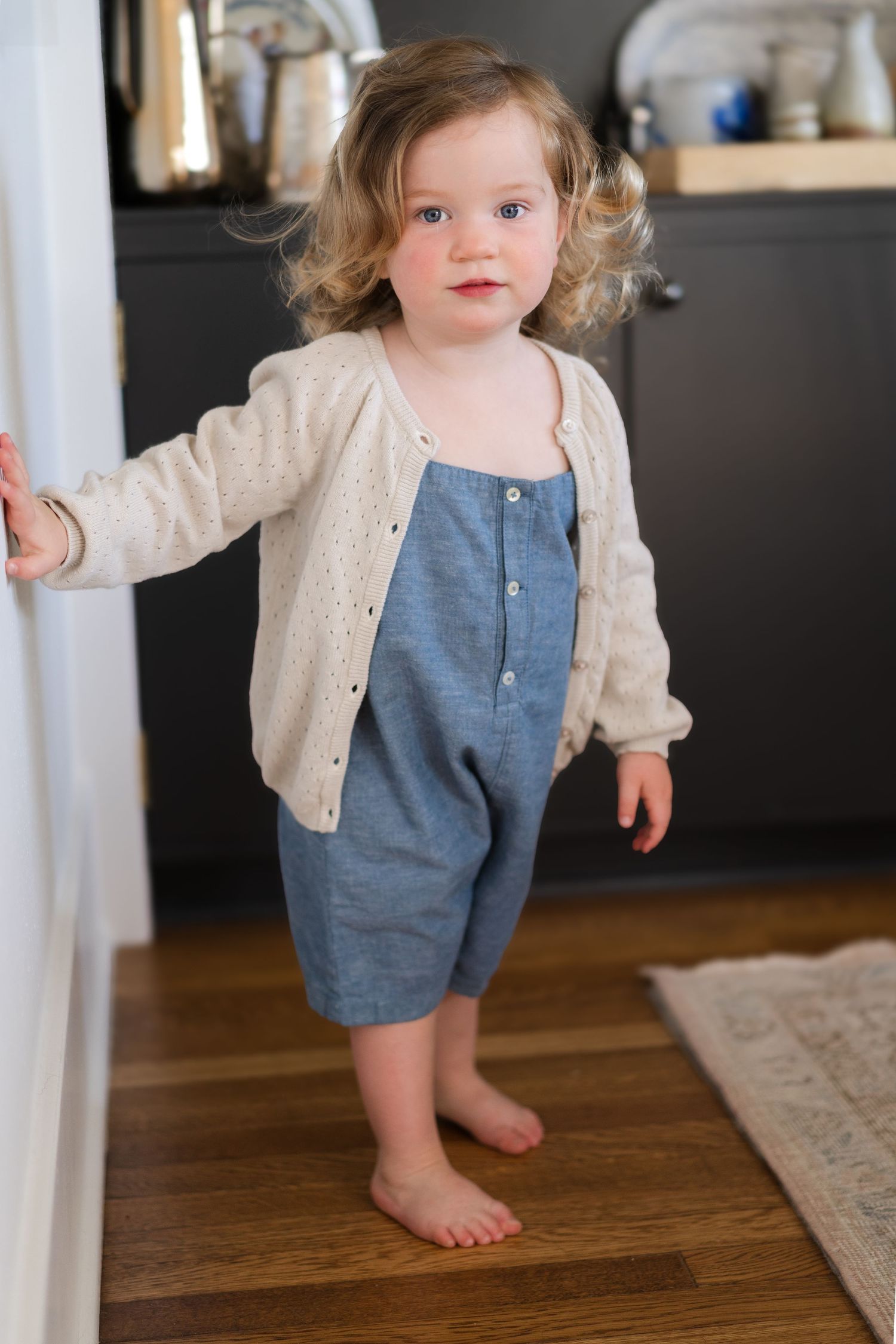 A toddler in denim overalls and cream cardigan stands barefoot on hardwood floors.
