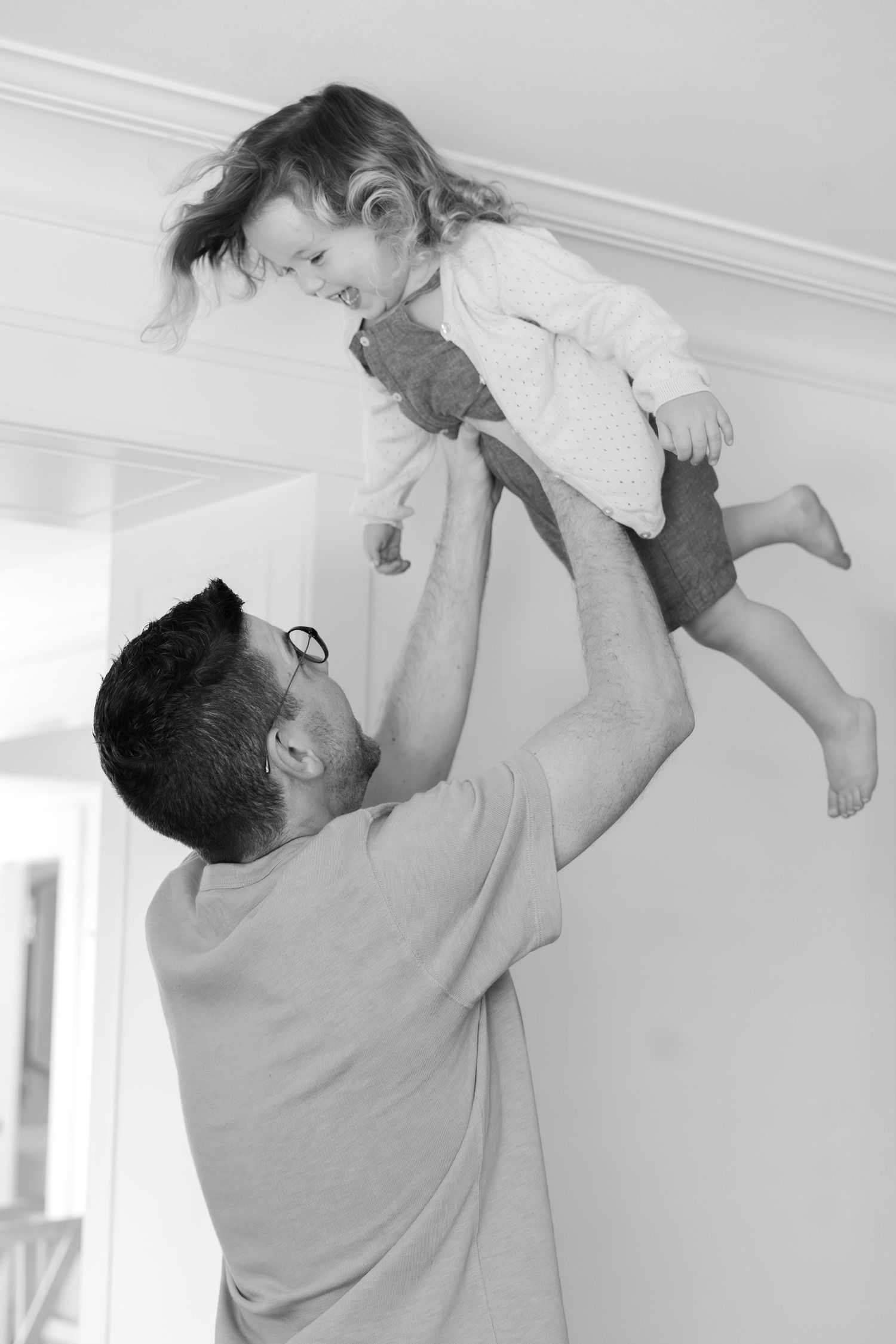 Playful black and white photo of parent lifting child in the air against white wall.