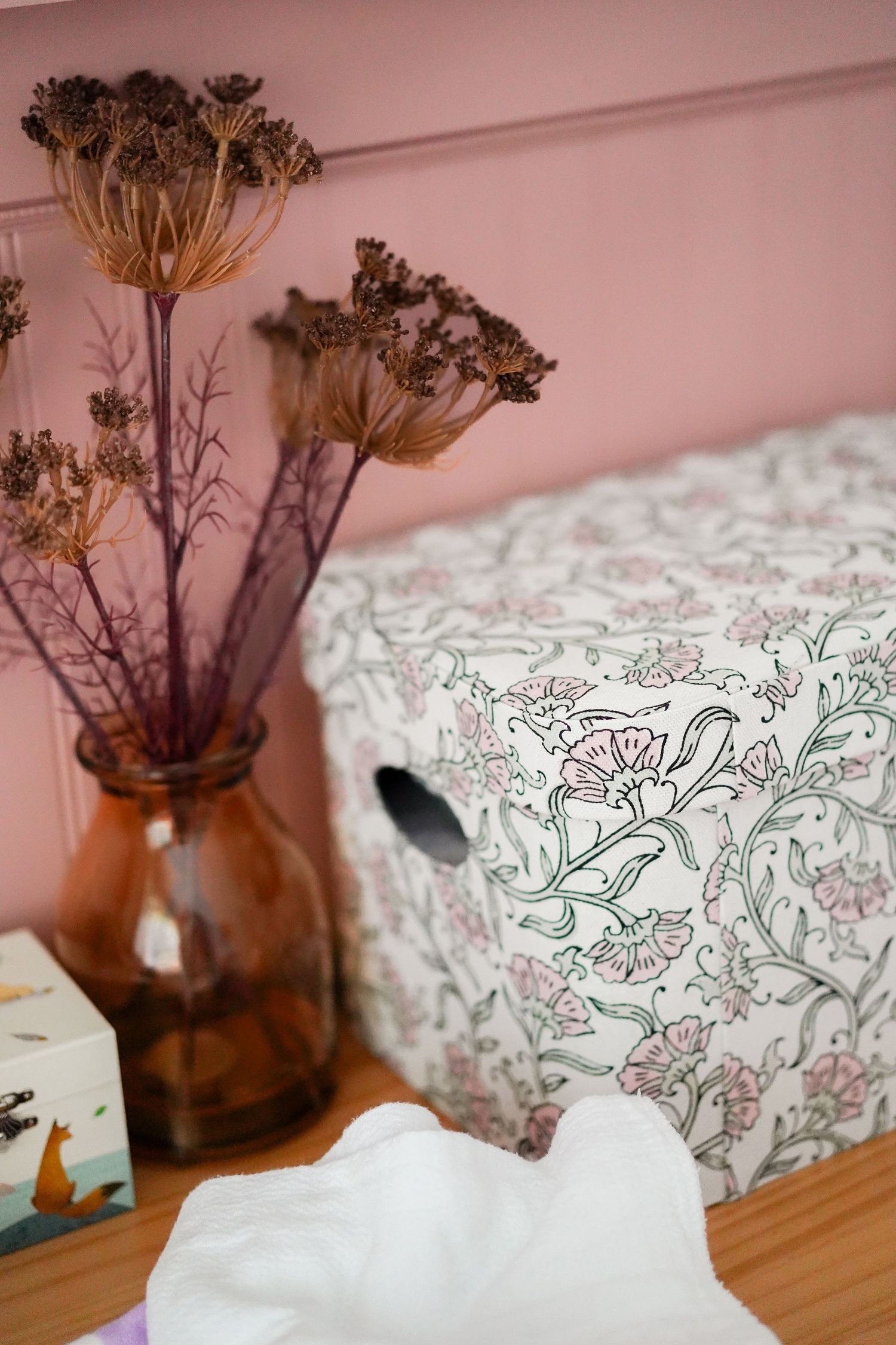 Dried flowers in amber vase next to patterned fabric on wooden surface.