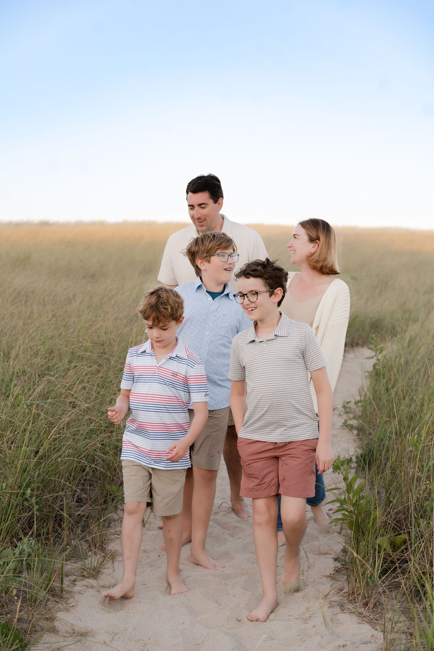 Family walking barefoot together through beach grass at sunset on a sandy coastal path.