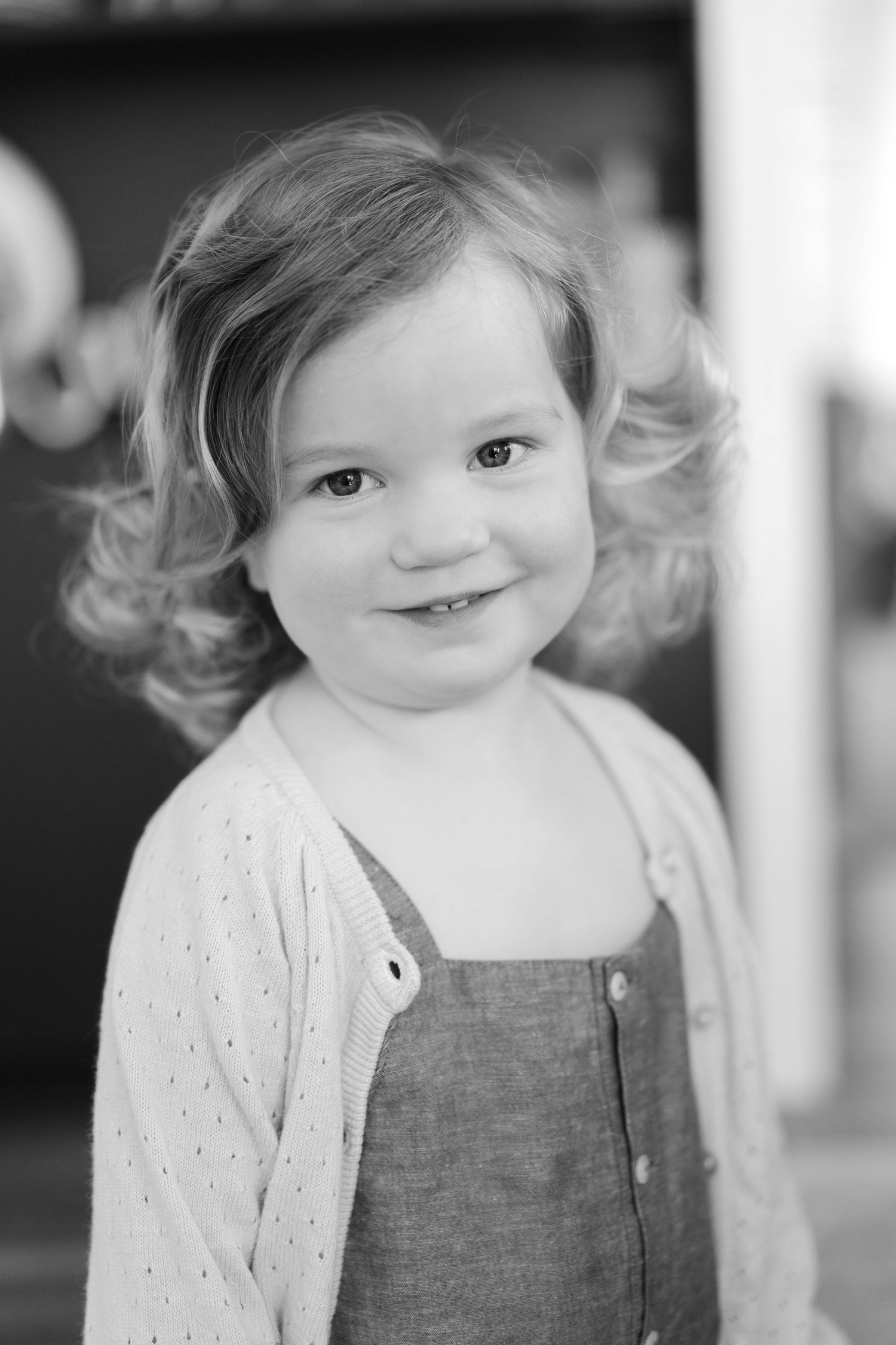 Child with curly hair poses in denim overalls and cream cardigan against dark background.