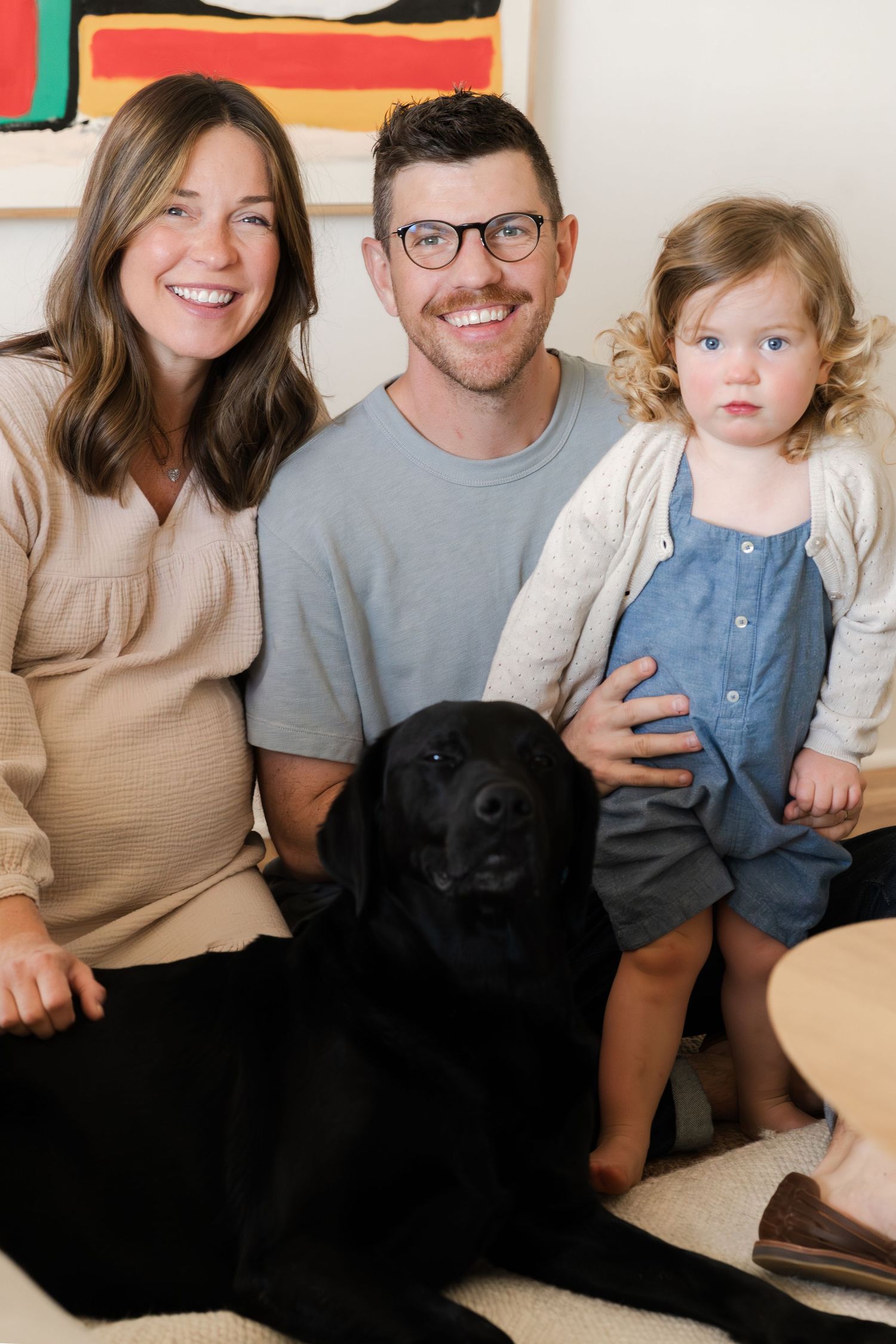 A casual family photo showing parents and child relaxing on couch with their black Labrador.