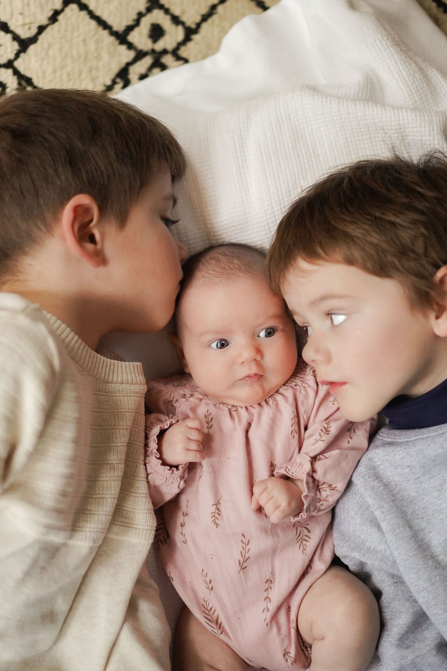 Three siblings sharing tender moment on white blanket with geometric rug visible.
