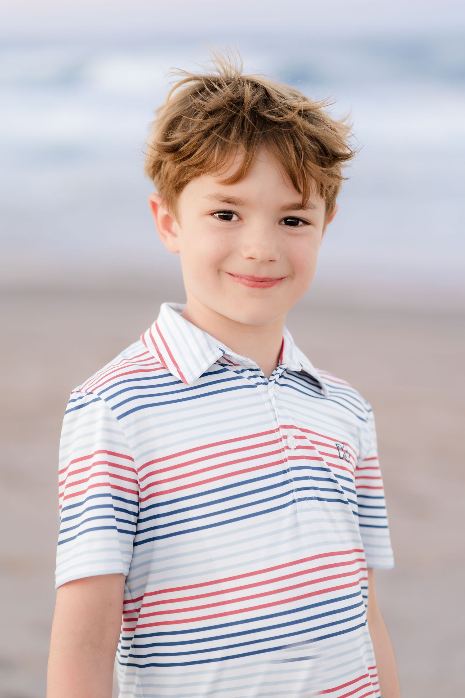 Child wearing a red and blue striped polo shirt smiles at the beach.
