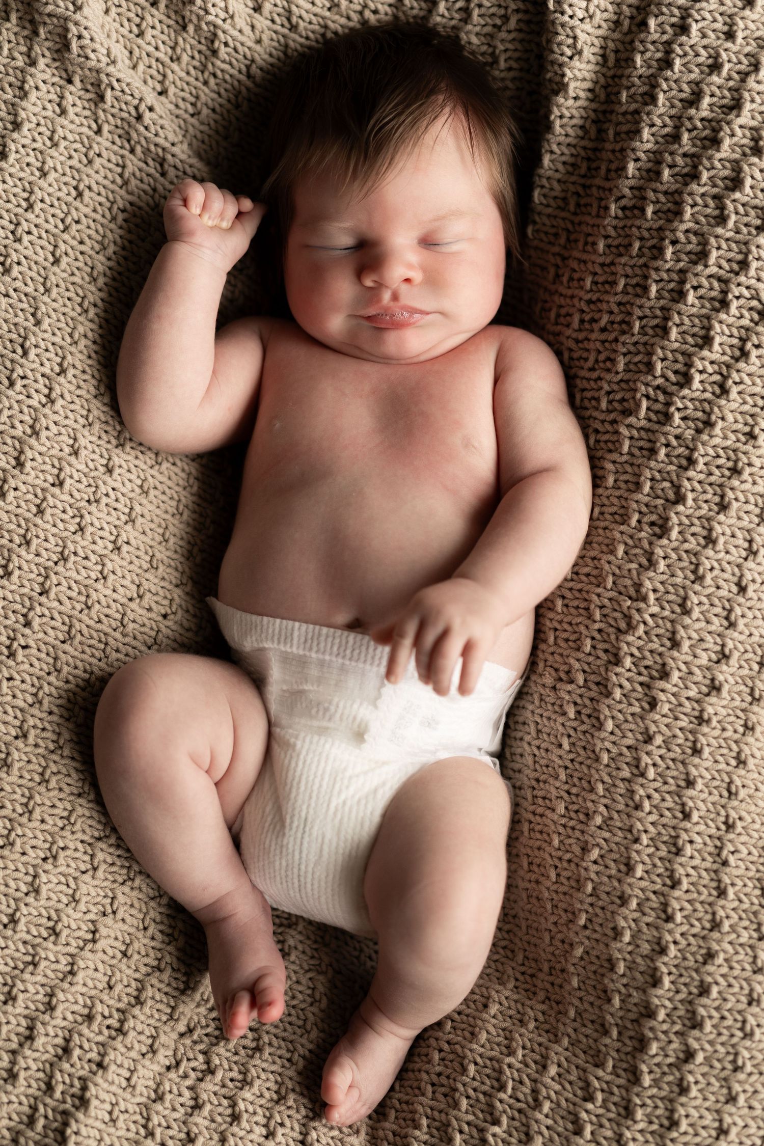Baby lying on textured beige knit blanket wearing white diaper in natural light photography.