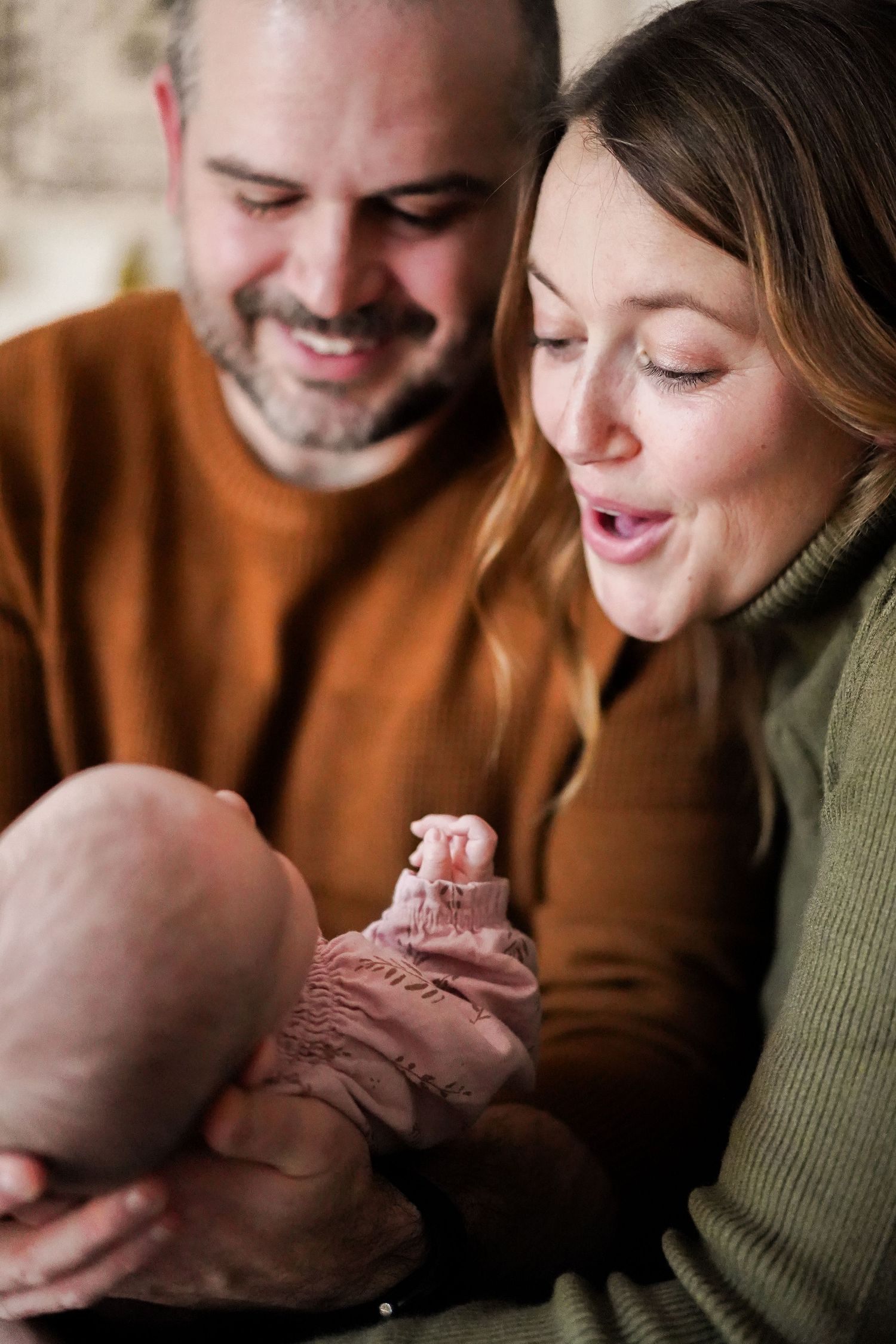 Intimate family portrait with warm lighting showing parents in earth-toned sweaters with baby.