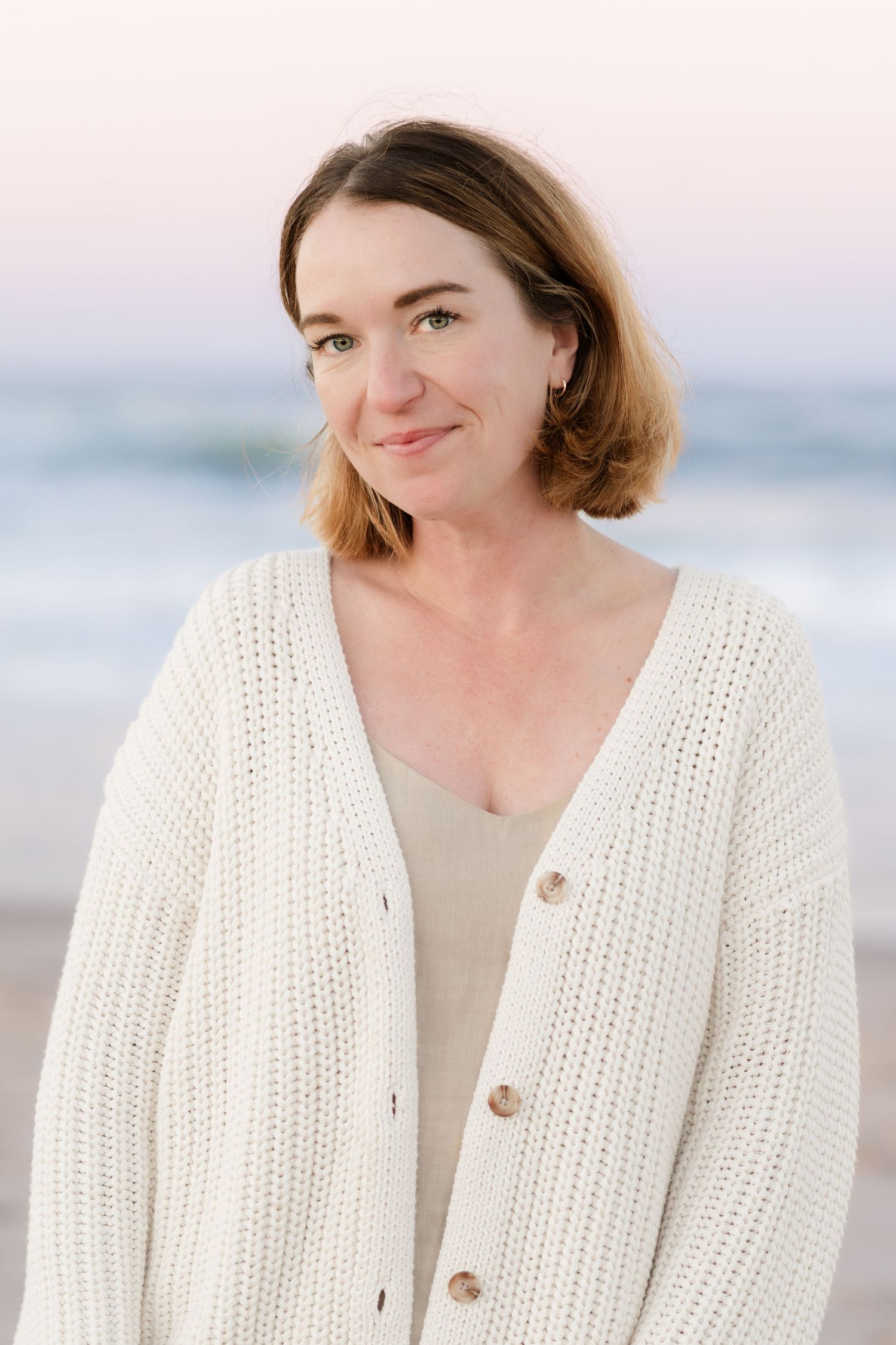 Woman in white knit cardigan and beige top poses at beach.