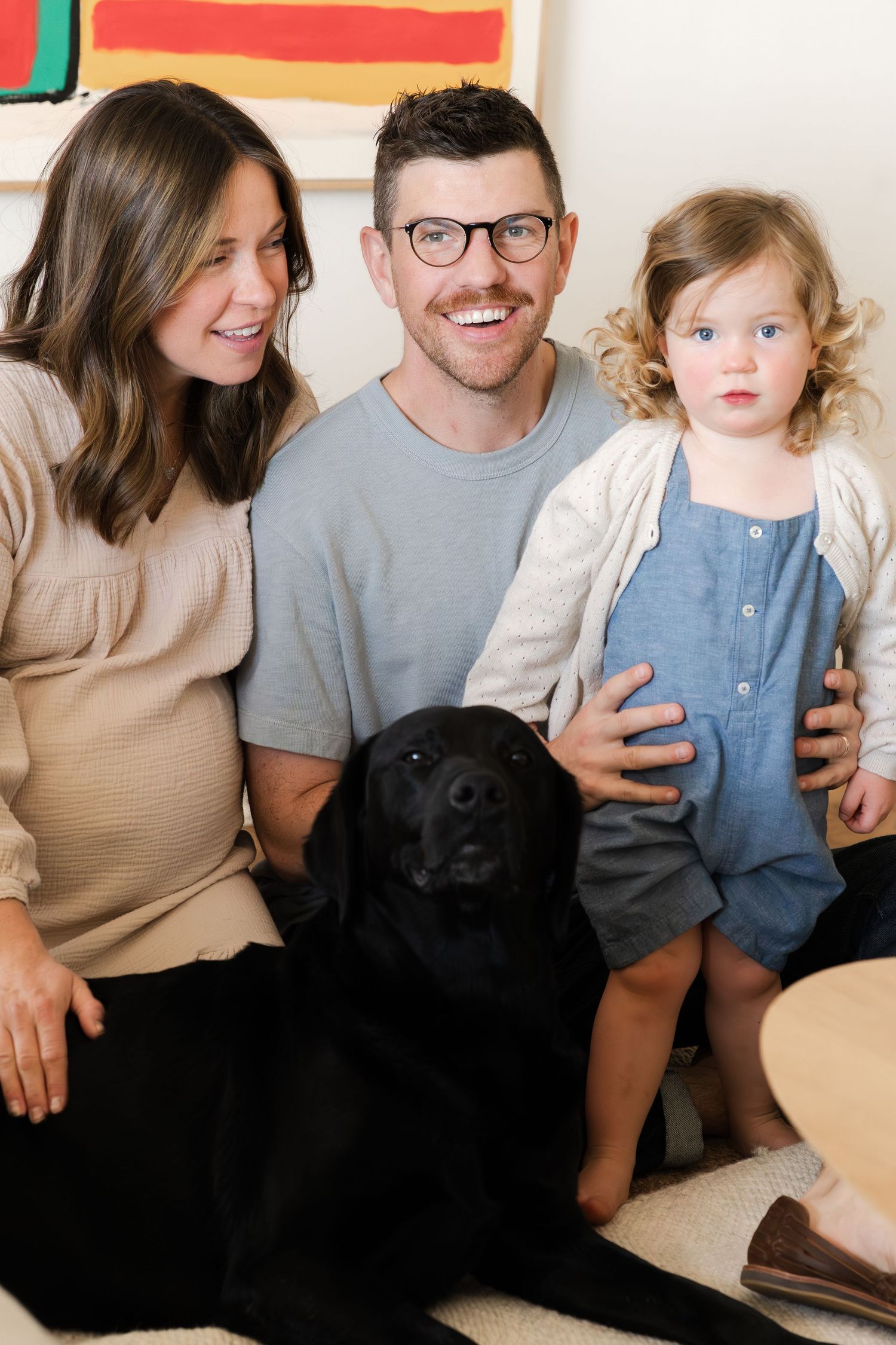 Family sitting together with their black dog on a couch against colorful wall art.