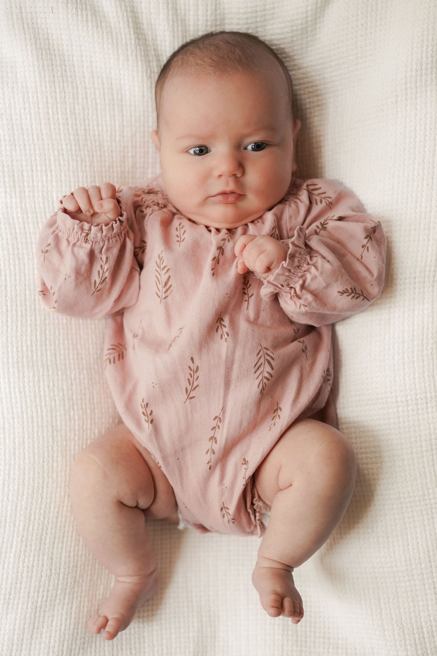 Baby wearing pink floral dress lying on white textured fabric.